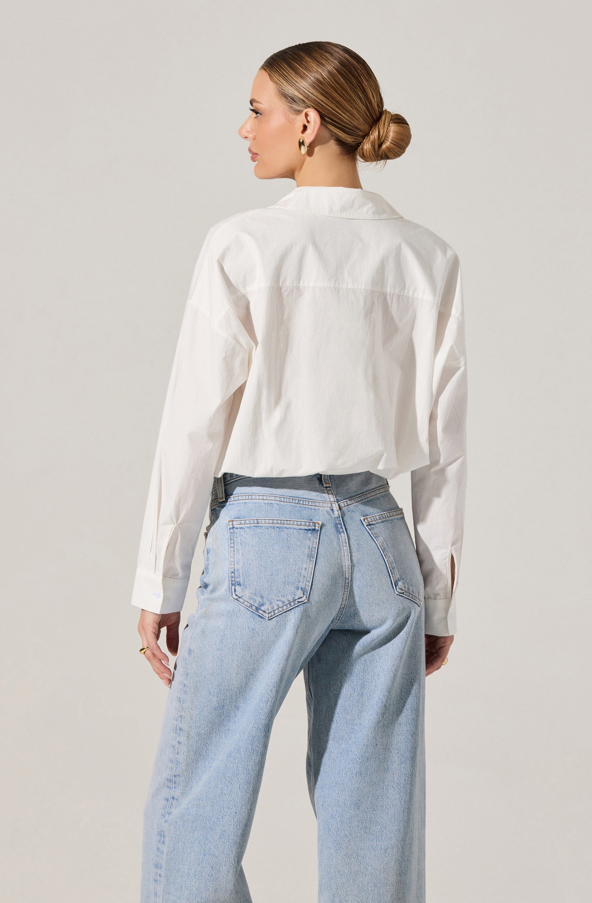 A woman is wearing a loose-fitting, long-sleeved white button-up shirt with a collar. The shirt has a slightly cropped length, ending just above the waist. She is also wearing light-wash, high-waisted jeans with a relaxed fit, featuring classic back pockets and visible stitching.