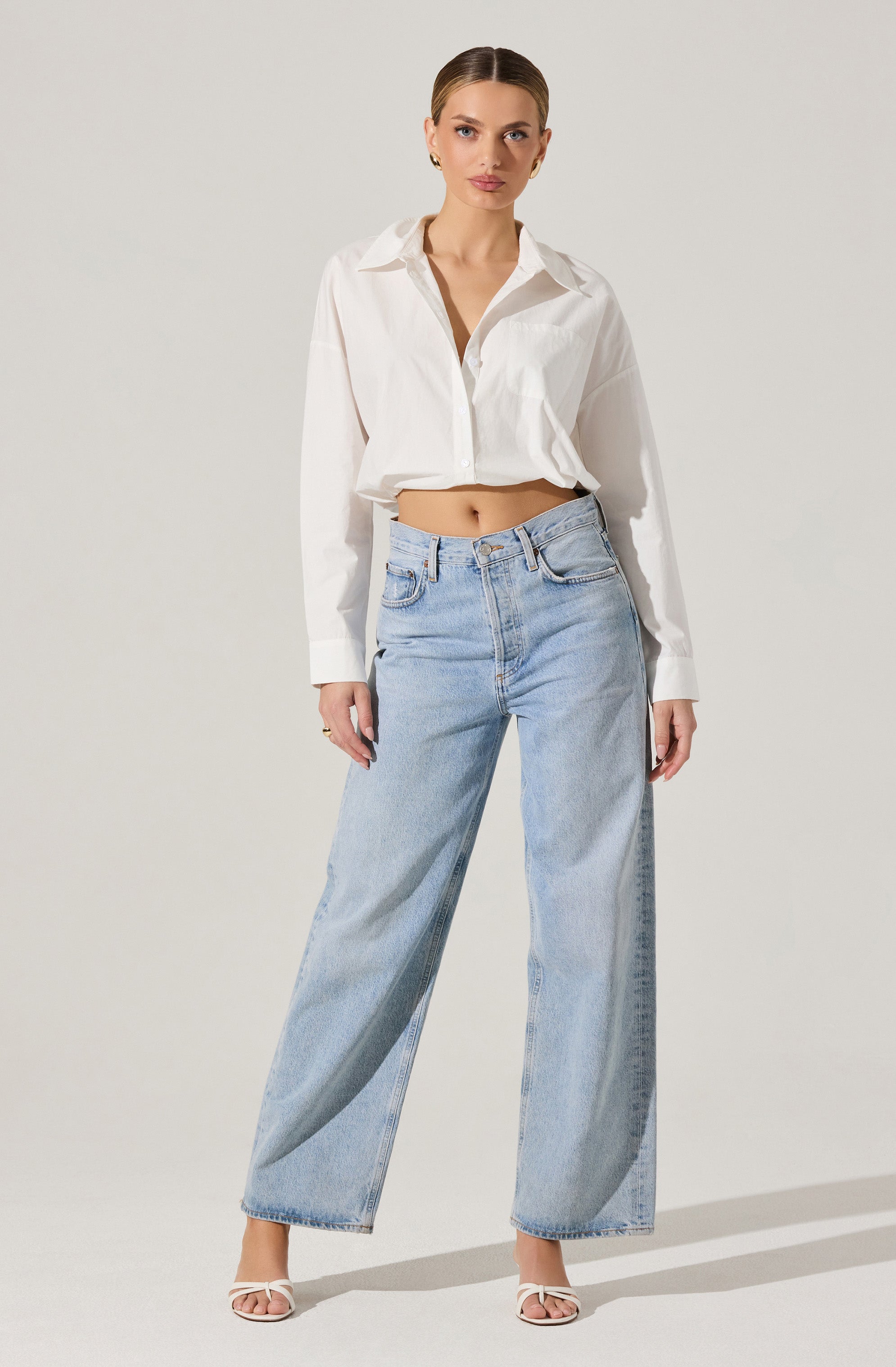 A woman is wearing an oversized white button-up shirt with a classic collar, long sleeves, and a slightly wrinkled texture. The shirt is unbuttoned at the top, revealing part of her midsection. She pairs the shirt with high-waisted, light-wash, wide-leg jeans that reach down to her ankles. She is also wearing open-toe, heeled sandals with thin white straps.