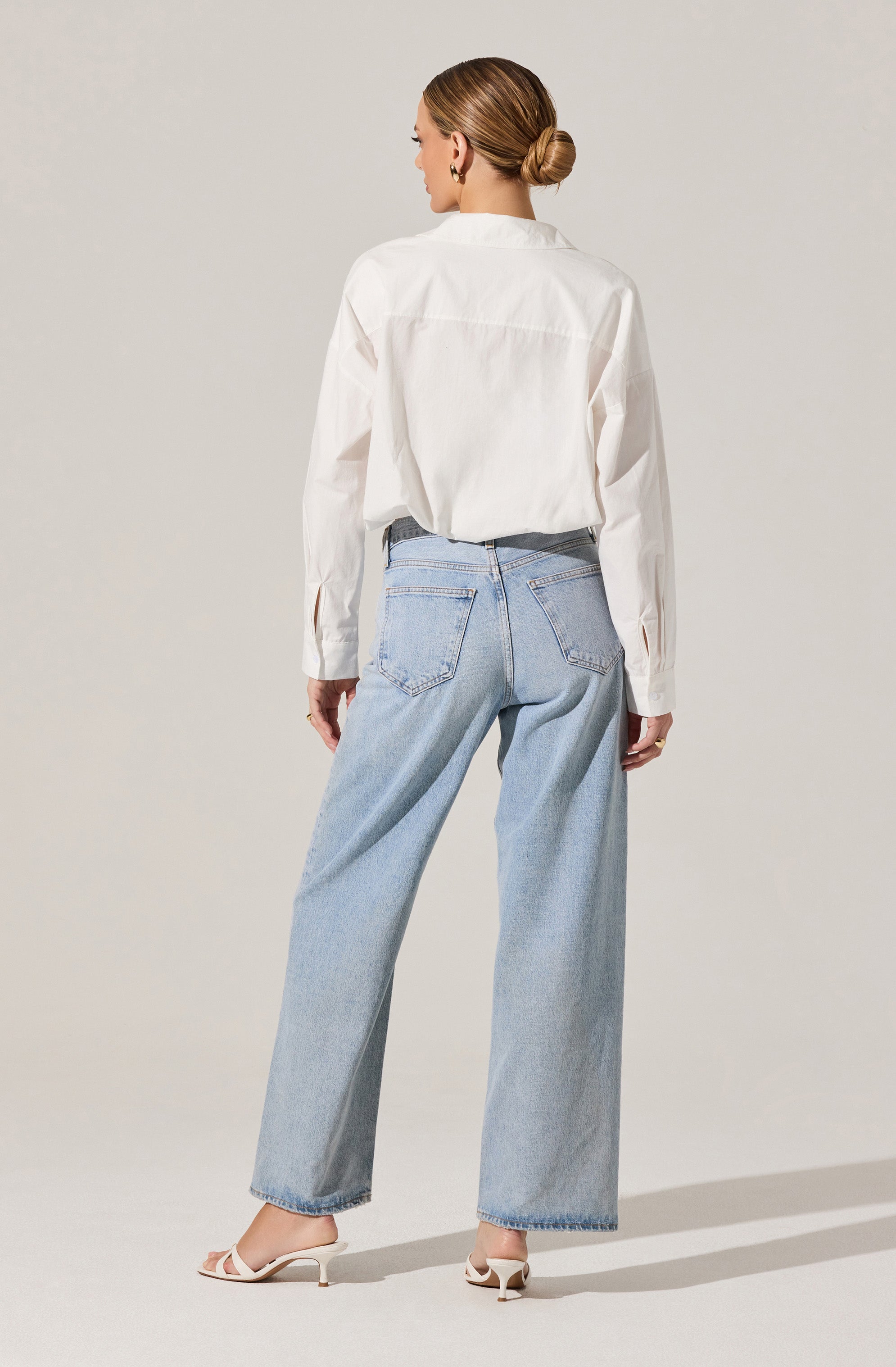 A woman is wearing a white long-sleeve button-up shirt with a loose fit, tucked into light-wash high-waisted jeans with a relaxed, wide-leg cut. She is also wearing white heeled sandals.