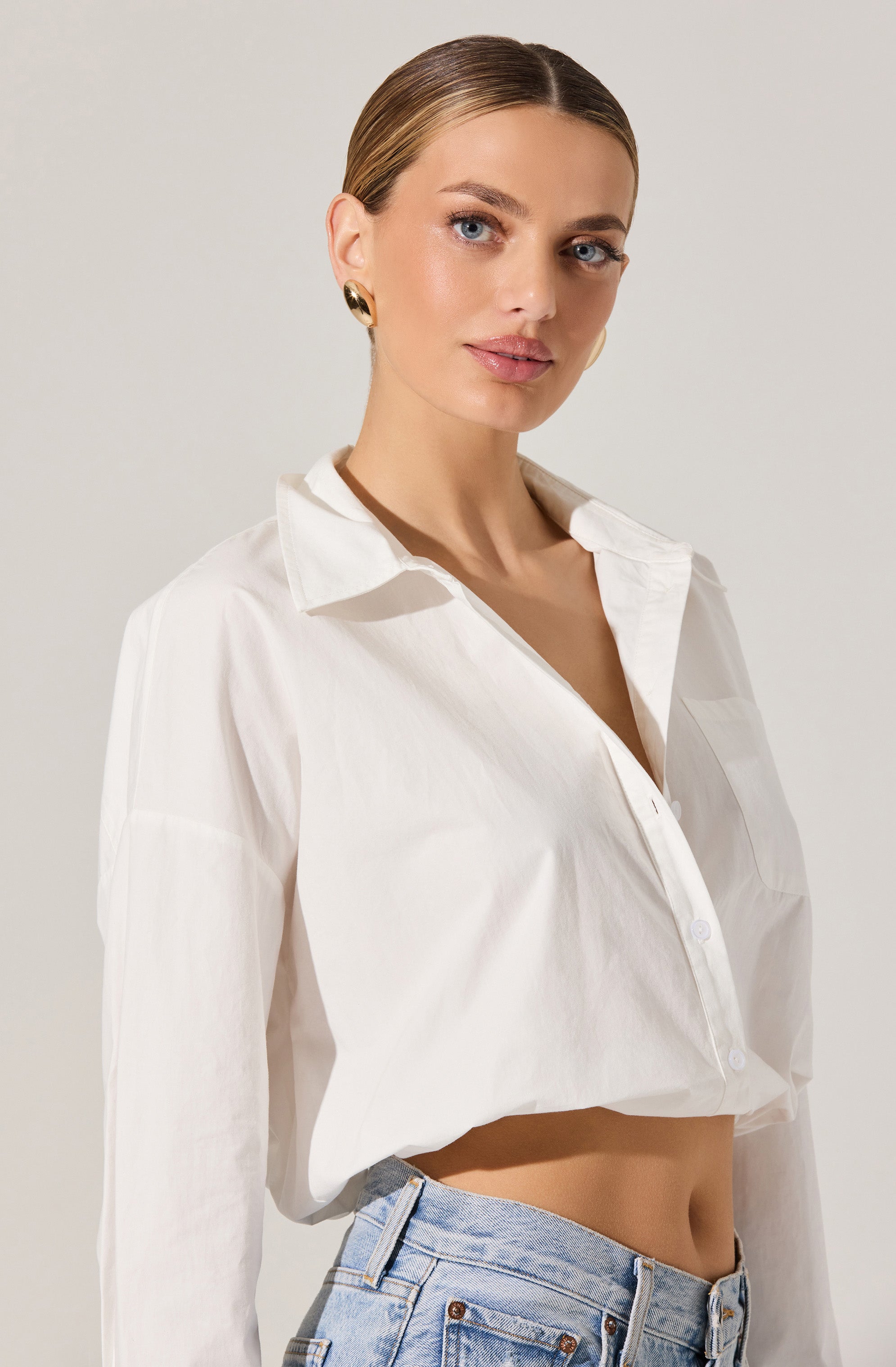 A woman is wearing a loose-fitting, long-sleeved white button-up shirt with a classic collar. The shirt is partially unbuttoned at the top, revealing her neckline. She is also wearing light blue high-waisted denim jeans.