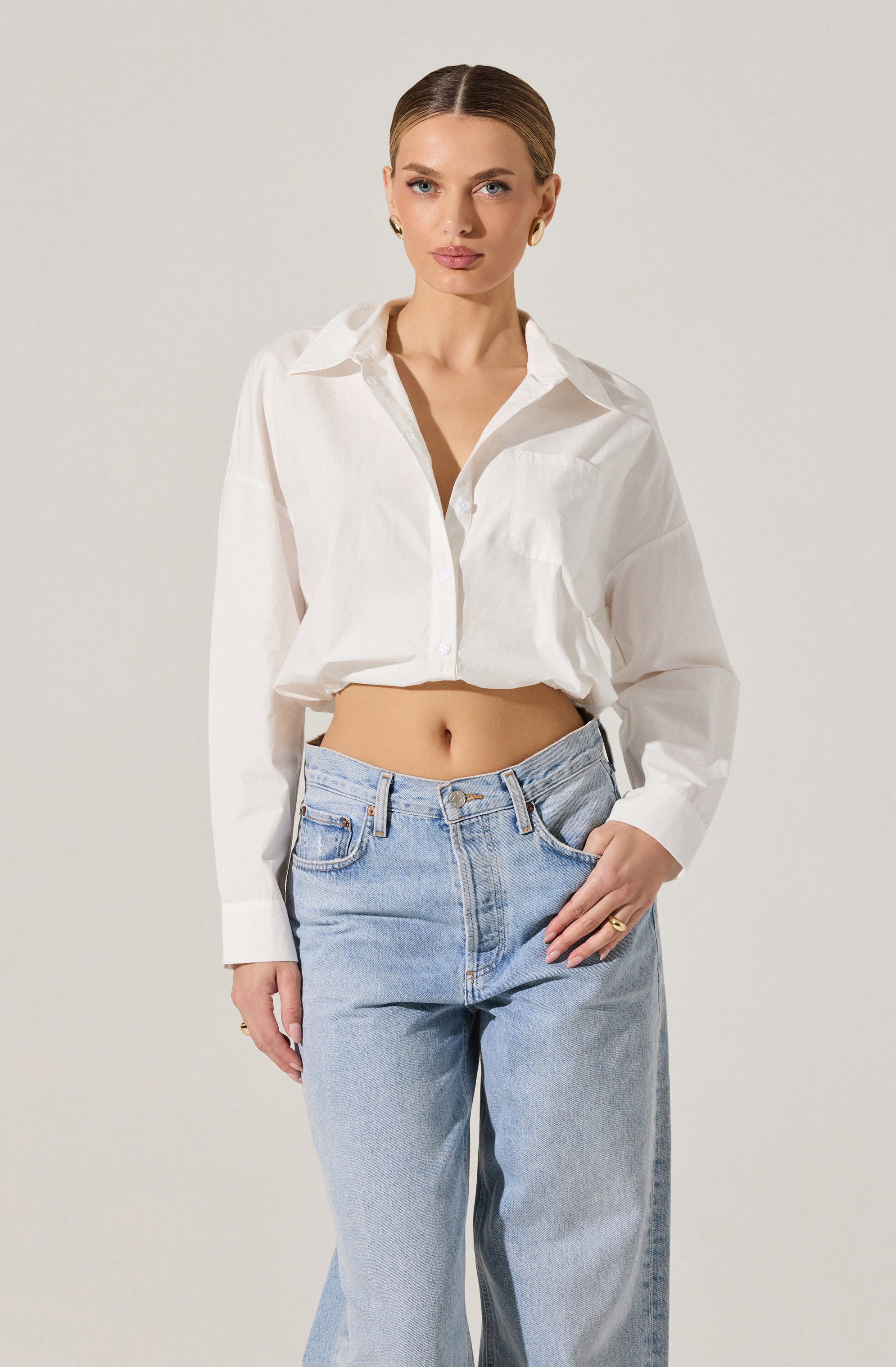 A woman is wearing a loose-fitting, long-sleeved white button-up shirt with a collar. The shirt is partly tucked into light blue, high-waisted jeans with a relaxed fit. The jeans have a classic five-pocket design and are fastened with a metal button.