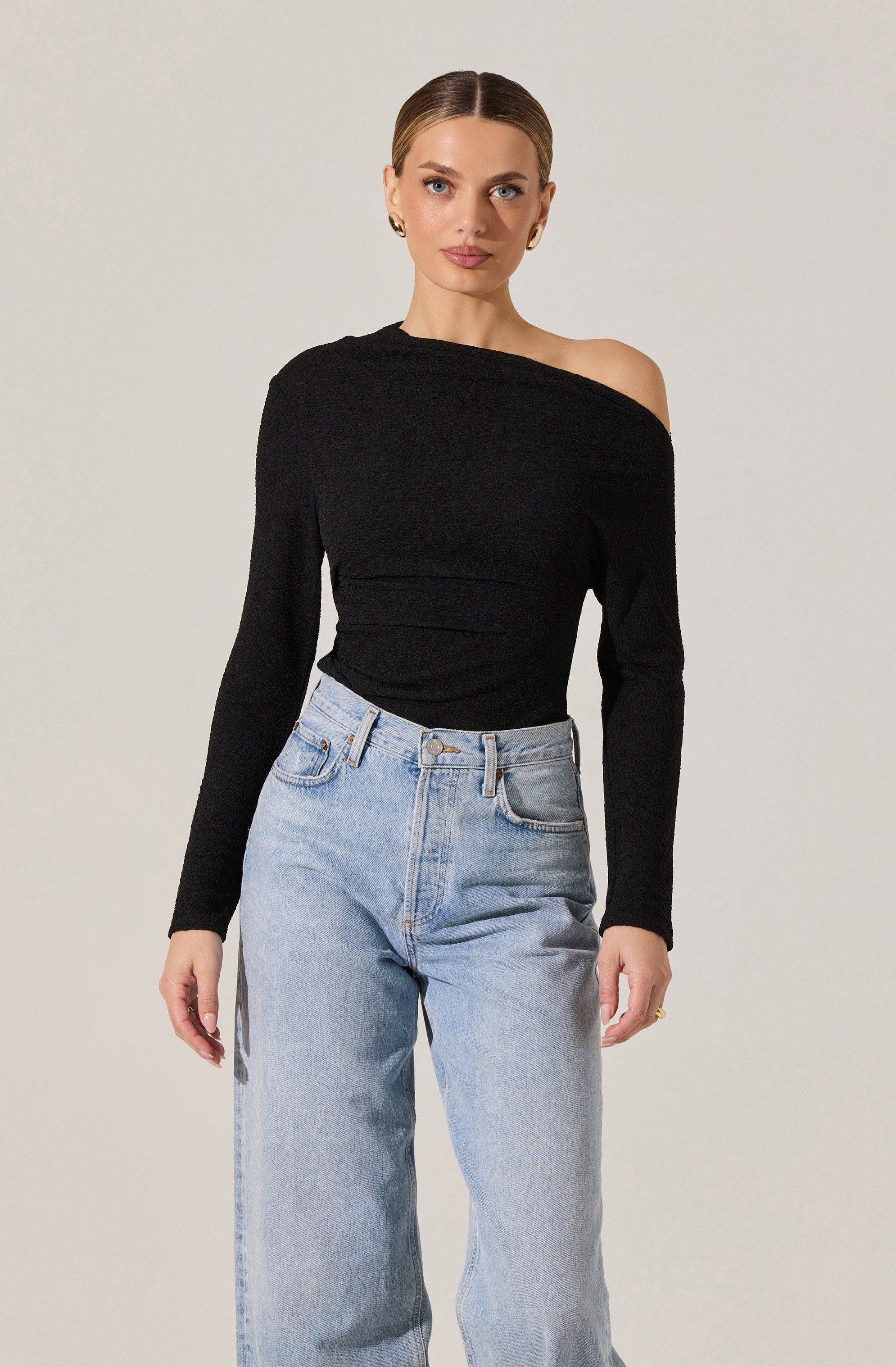 A woman is wearing a black, off-the-shoulder long-sleeve top paired with light-wash, high-waisted jeans. The top has a textured knit appearance and covers one shoulder while exposing the other. The jeans have a classic five-pocket design.
