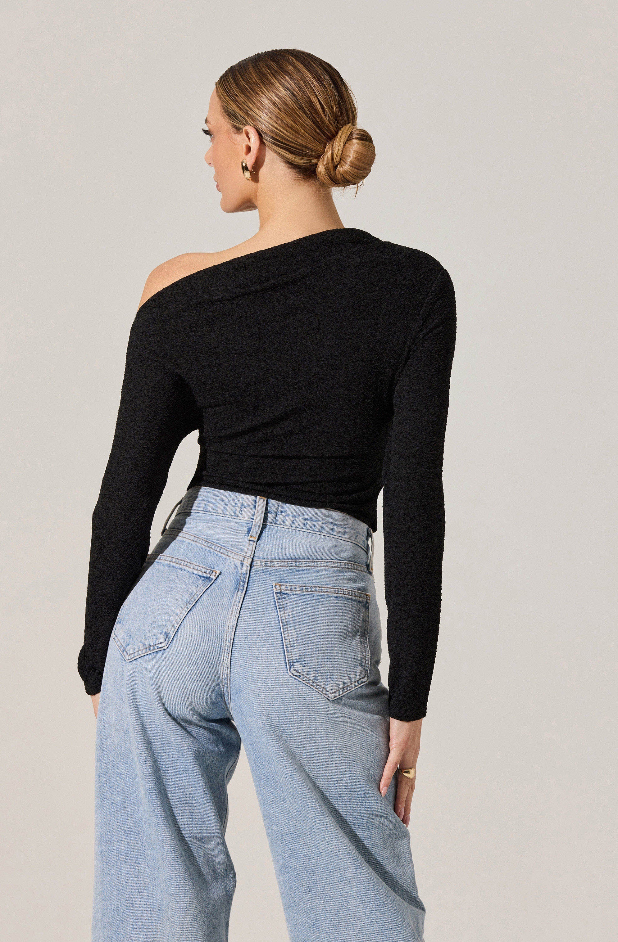 A woman is wearing a black, long-sleeved, textured top that features an off-the-shoulder design on one side. She pairs it with high-waisted, light-wash denim jeans with a relaxed fit, featuring back pockets and visible stitching.