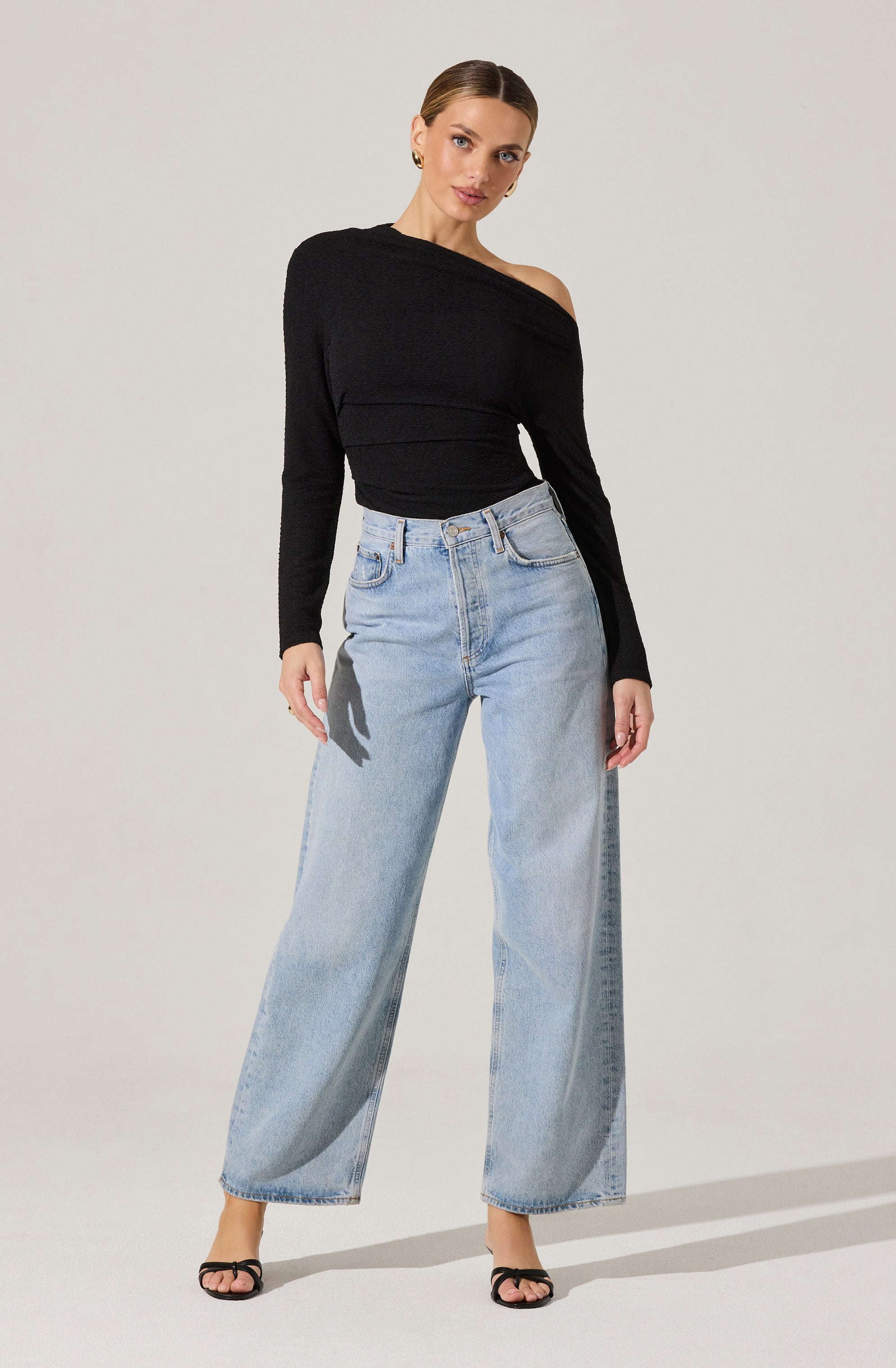 The woman is wearing a black, off-the-shoulder sweater with a loose fit, featuring long sleeves and a layered, textured design across the chest. She pairs it with high-waisted, light-wash wide-leg jeans that have a relaxed fit and a classic five-pocket style. She completes the outfit with black open-toe heels.