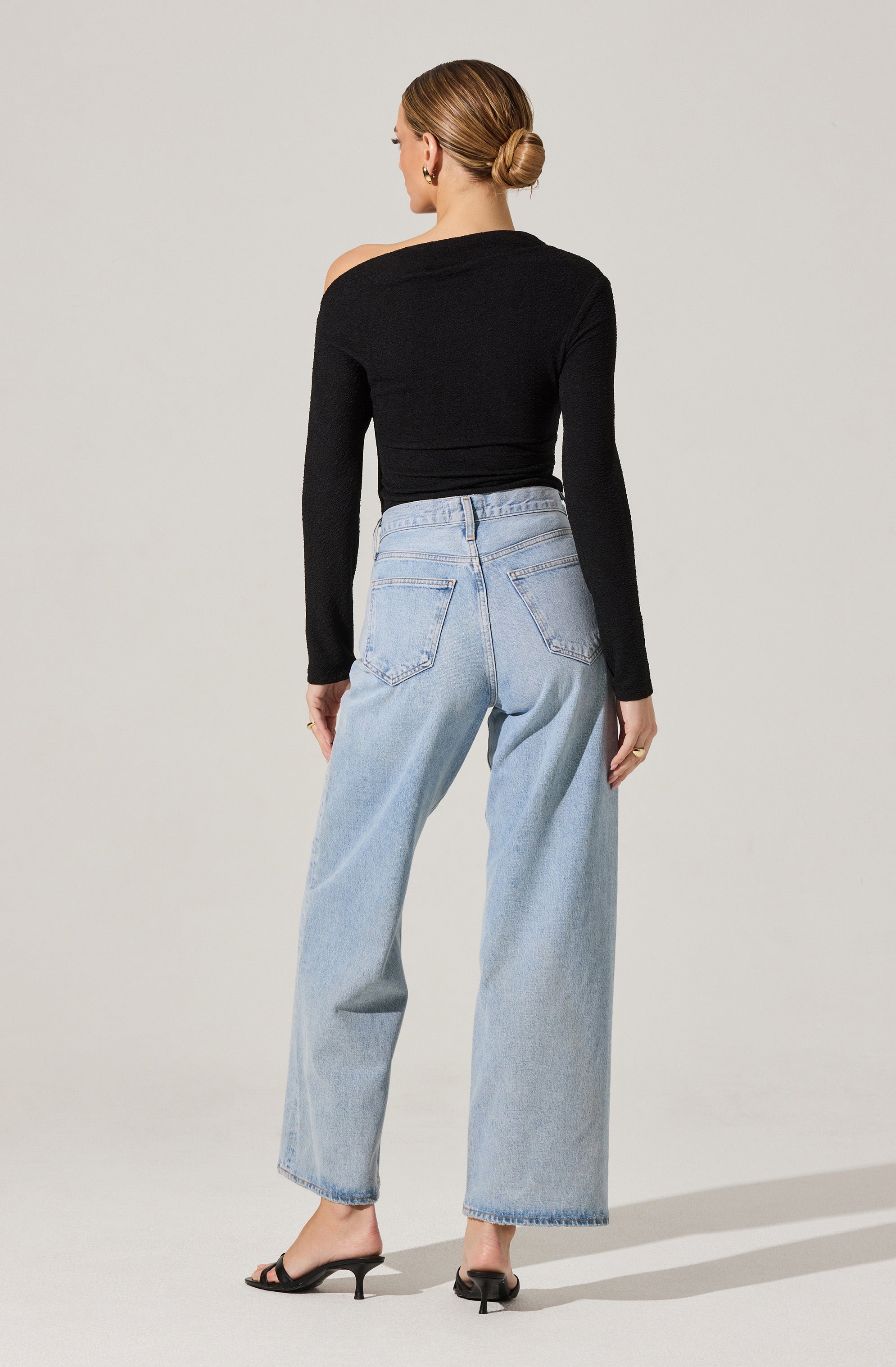A woman is wearing a fitted black long-sleeve top with an asymmetrical shoulder design, paired with high-waisted, light-wash wide-leg jeans featuring large back pockets. She is also wearing black open-toe heeled sandals.