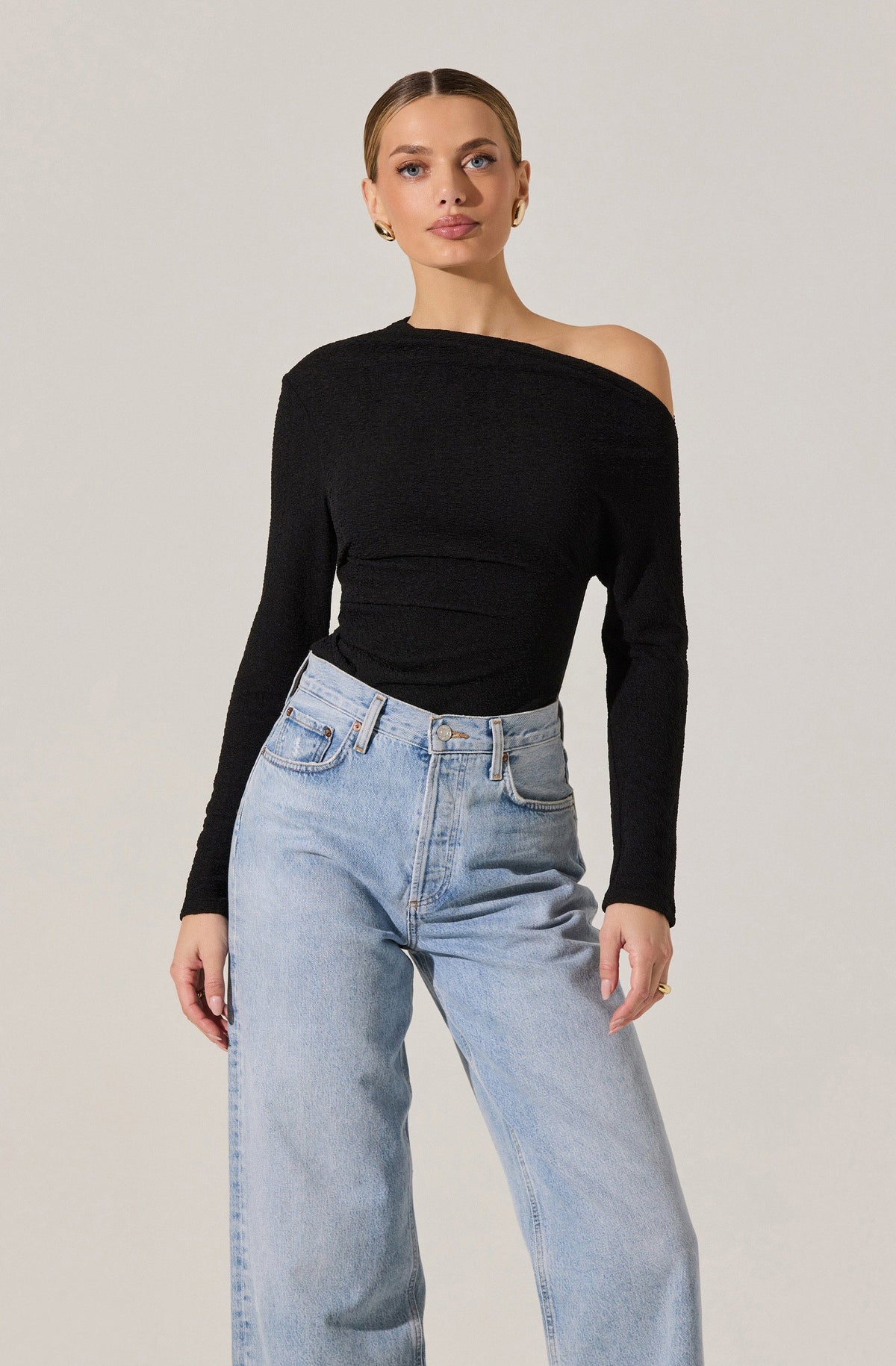 A woman is wearing a black, long-sleeved, off-the-shoulder top with a textured fabric, paired with high-waisted, light-wash jeans.