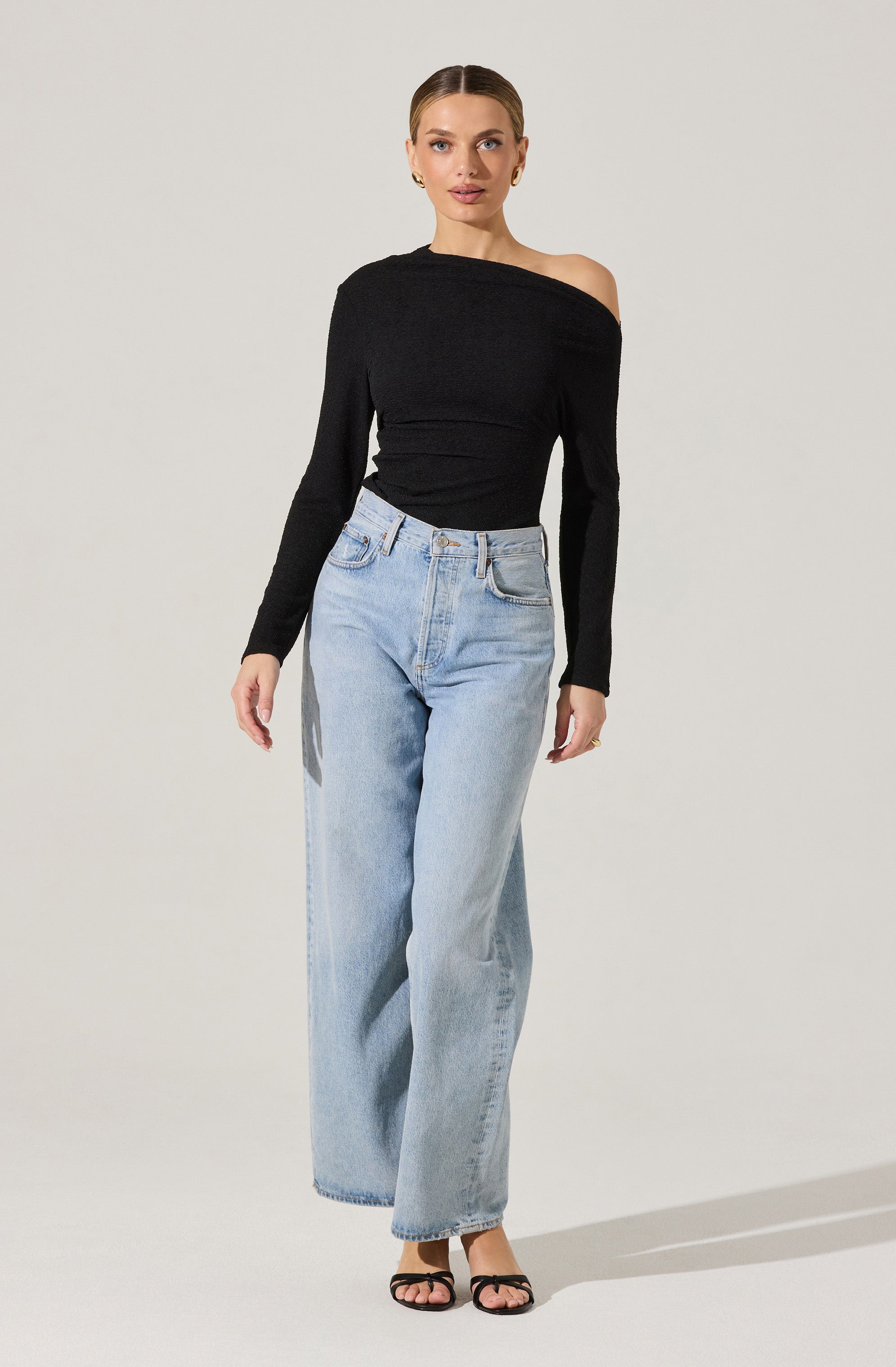 A woman is wearing a black, off-the-shoulder long-sleeve top paired with light-wash, high-waisted wide-leg jeans. She is also wearing black strappy high-heeled sandals.