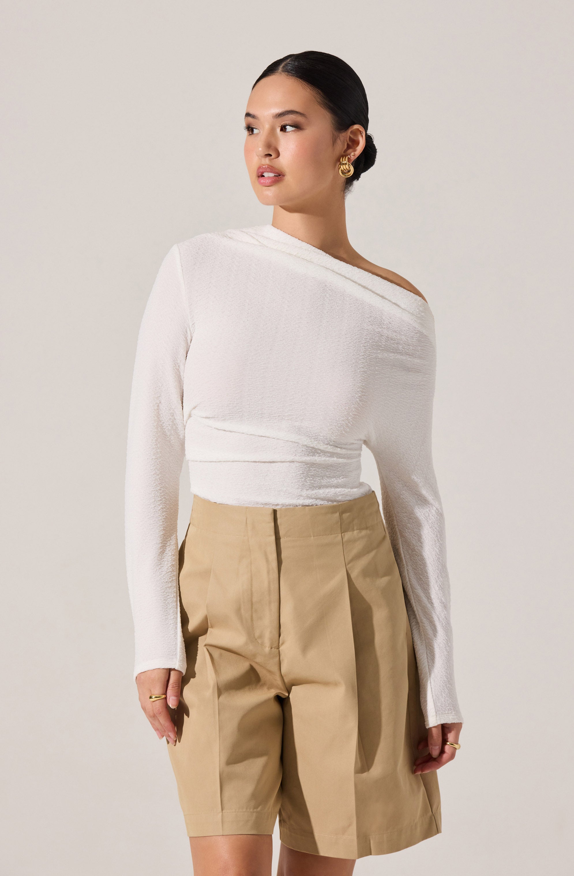 A woman is wearing a white, long-sleeved knit top with an off-the-shoulder neckline, paired with high-waisted beige shorts that have pleats and a front zipper.