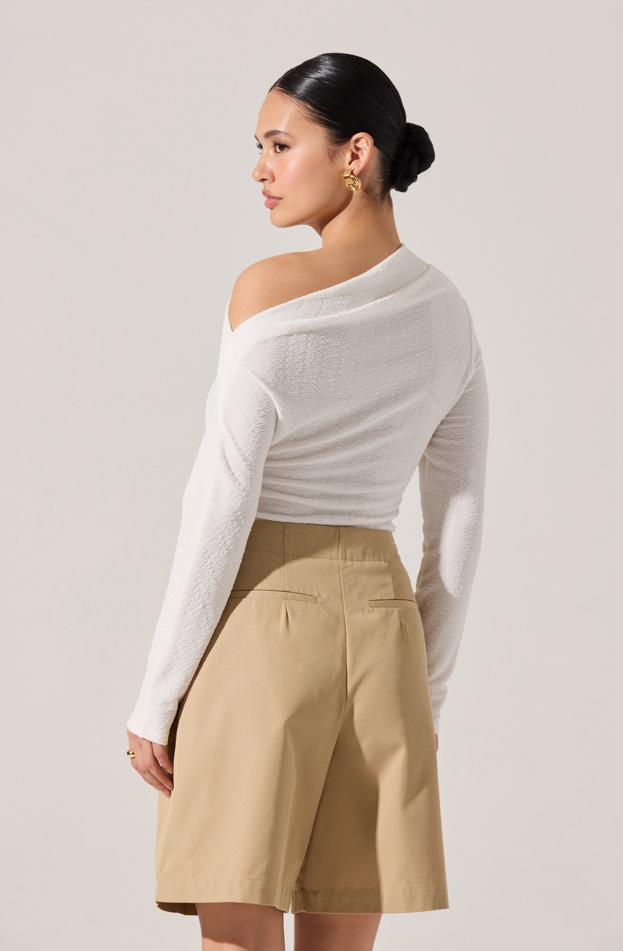 A woman is wearing a white, long-sleeved, off-the-shoulder sweater with a textured fabric, paired with high-waisted beige shorts that have a tailored fit and back welt pockets.