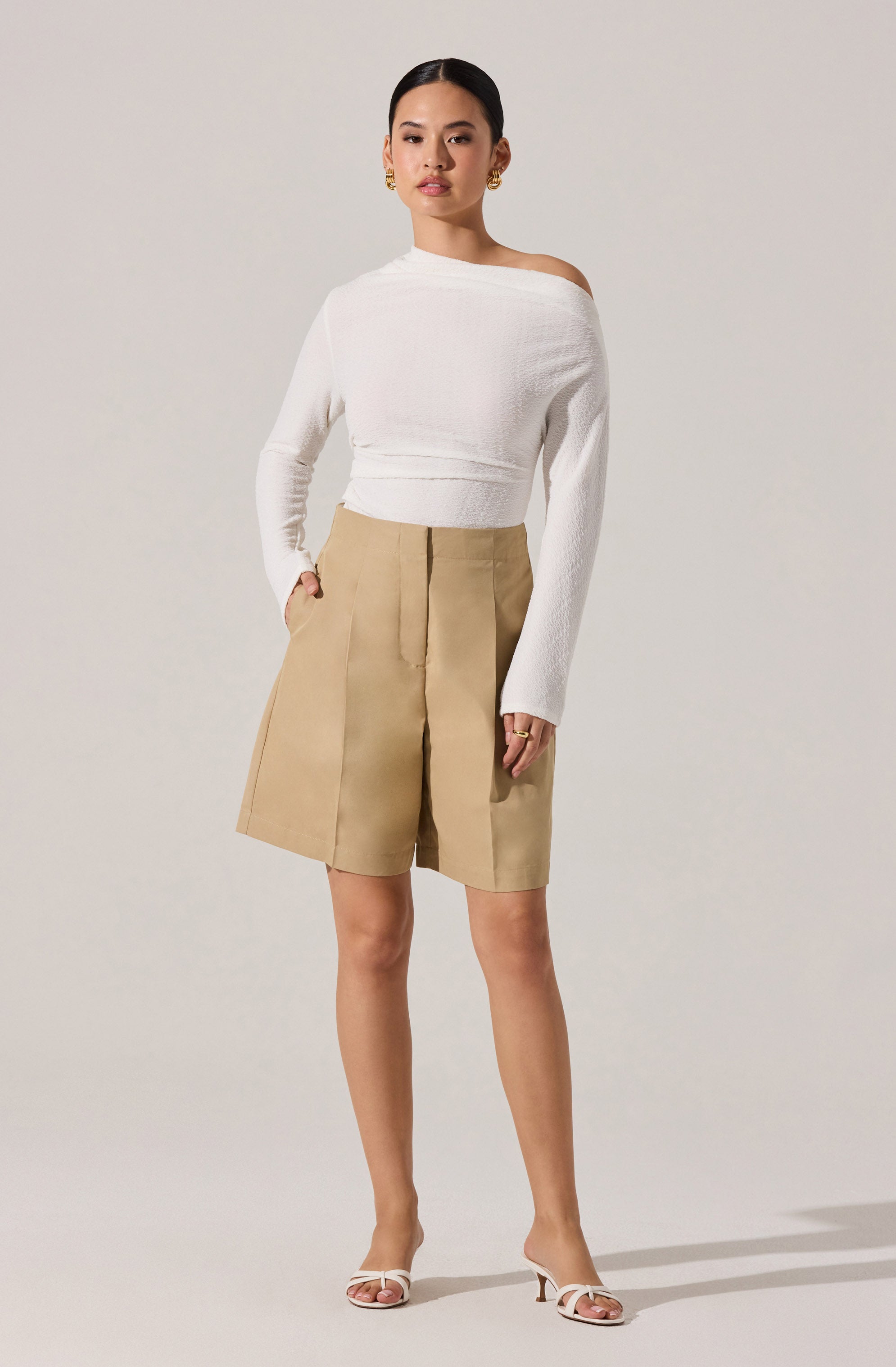 The woman is wearing a white, long-sleeved, off-the-shoulder sweater with a textured knit pattern. She pairs it with high-waisted beige shorts that have a wide-leg fit and front pleats. She completes her outfit with white, open-toe high-heeled sandals.