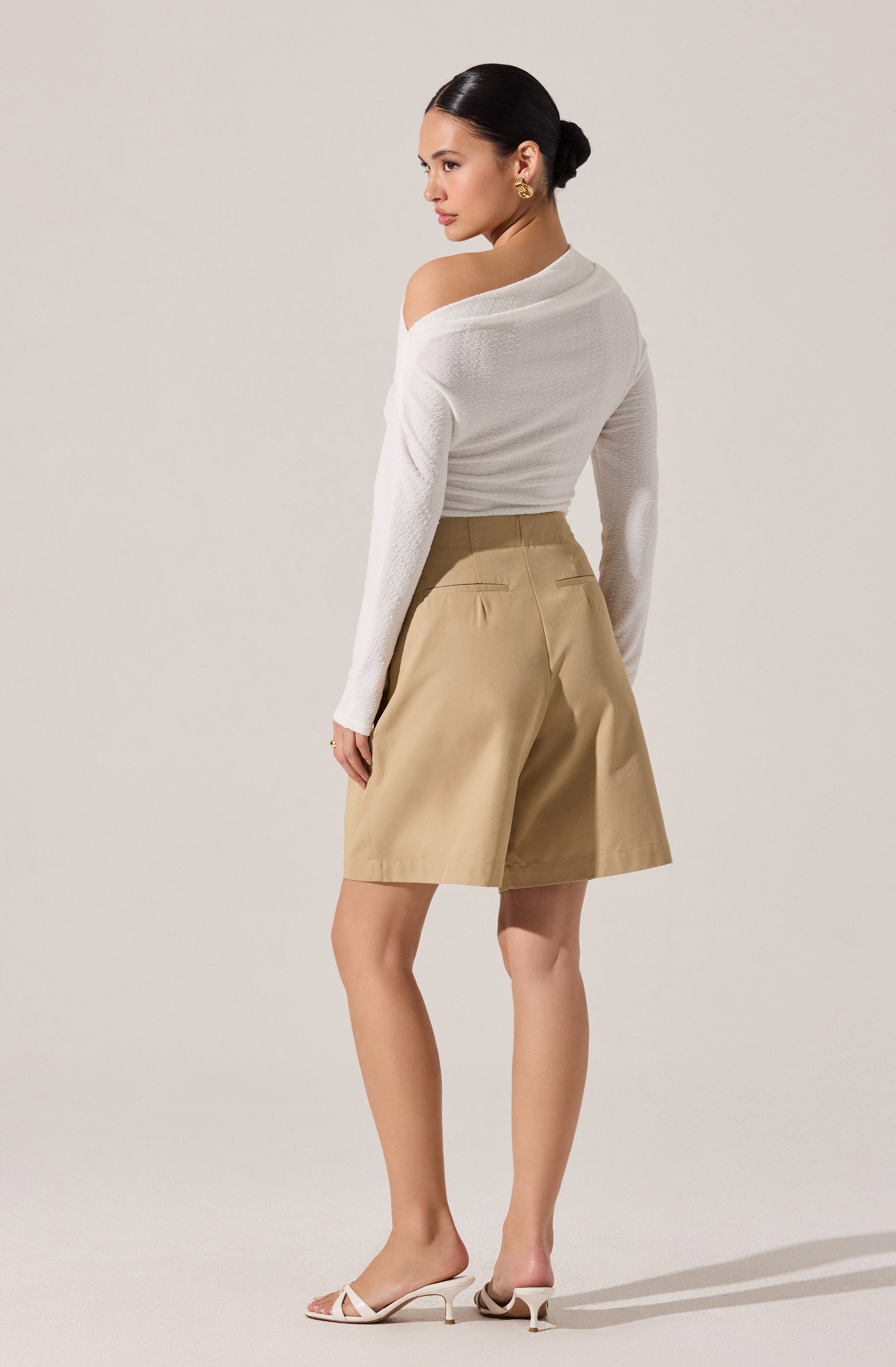 A woman is wearing a white, long-sleeved, off-the-shoulder sweater with a textured pattern. She has on high-waisted, khaki-colored shorts with back pockets. She accessorizes with gold earrings and is wearing beige open-toe high-heeled sandals. Her hair is styled in a sleek, low bun.