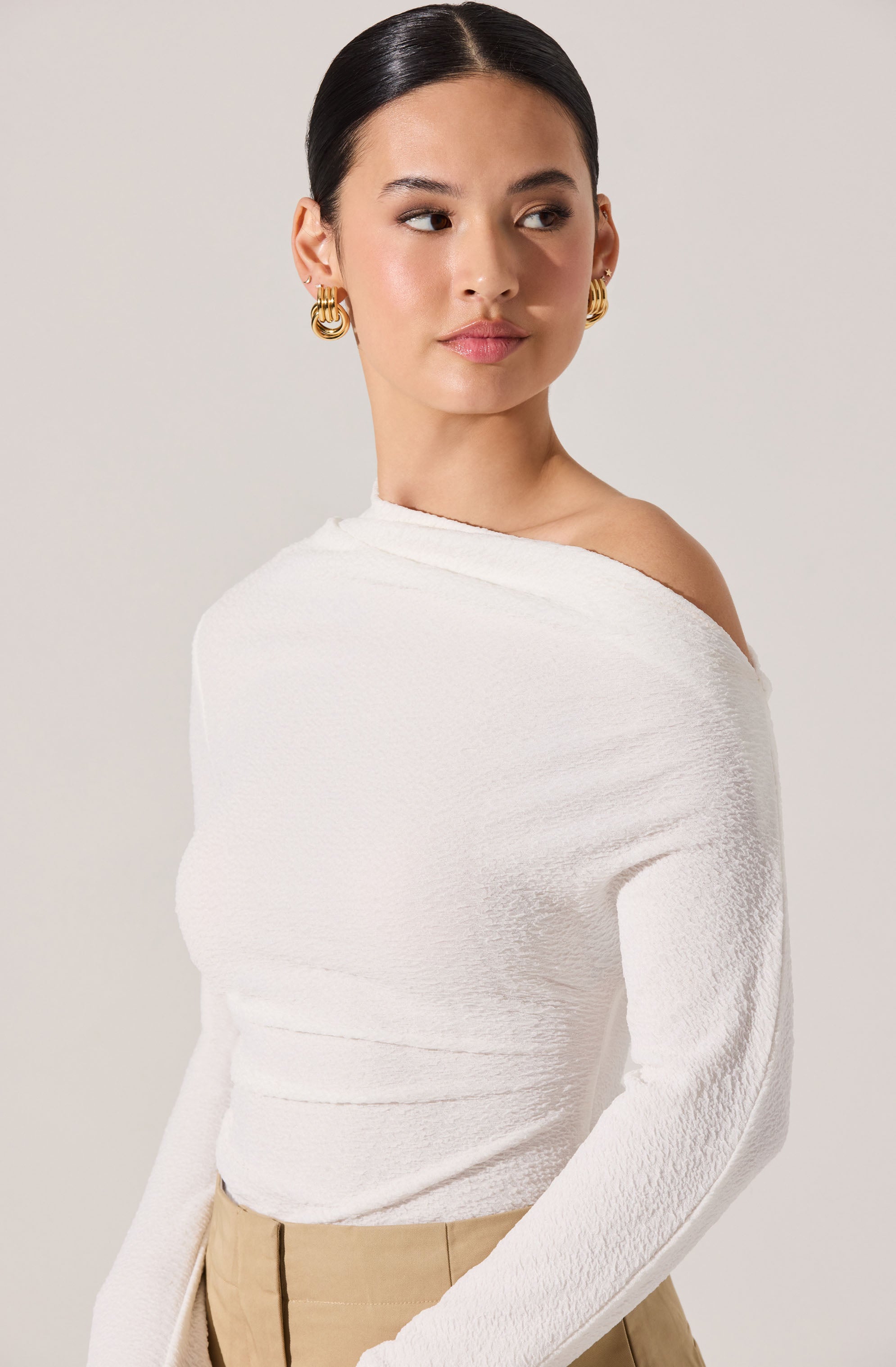 A woman with dark hair styled in a sleek, low bun is wearing a white, textured, off-the-shoulder top that exposes one shoulder. She has on gold hoop earrings and beige high-waisted pants.