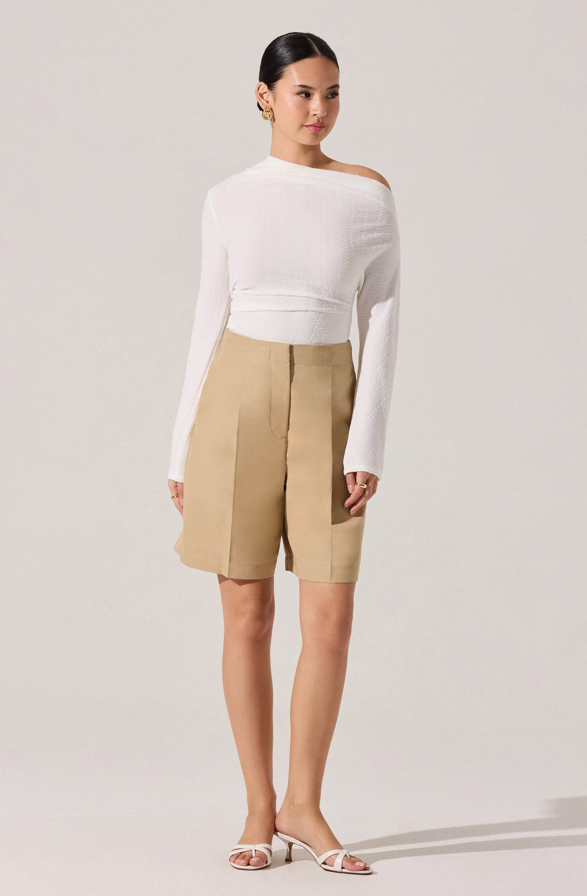 A woman is wearing a white, long-sleeved knit sweater with an off-the-shoulder design on one side, paired with high-waisted beige shorts that feature wide pleats and a large patch pocket on the right side. She is accessorized with gold earrings and rings, and is wearing white strappy heeled sandals.