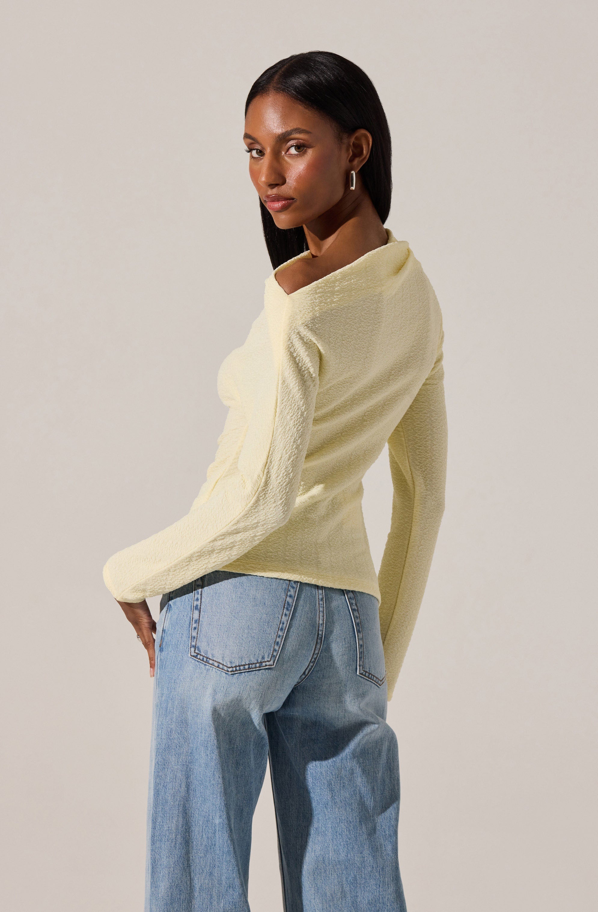 A woman with dark skin and straight, shoulder-length black hair is wearing a light yellow textured long-sleeve top with a wide neckline, paired with high-waisted, loose-fitting light blue jeans. She is also accessorized with small hoop earrings.
