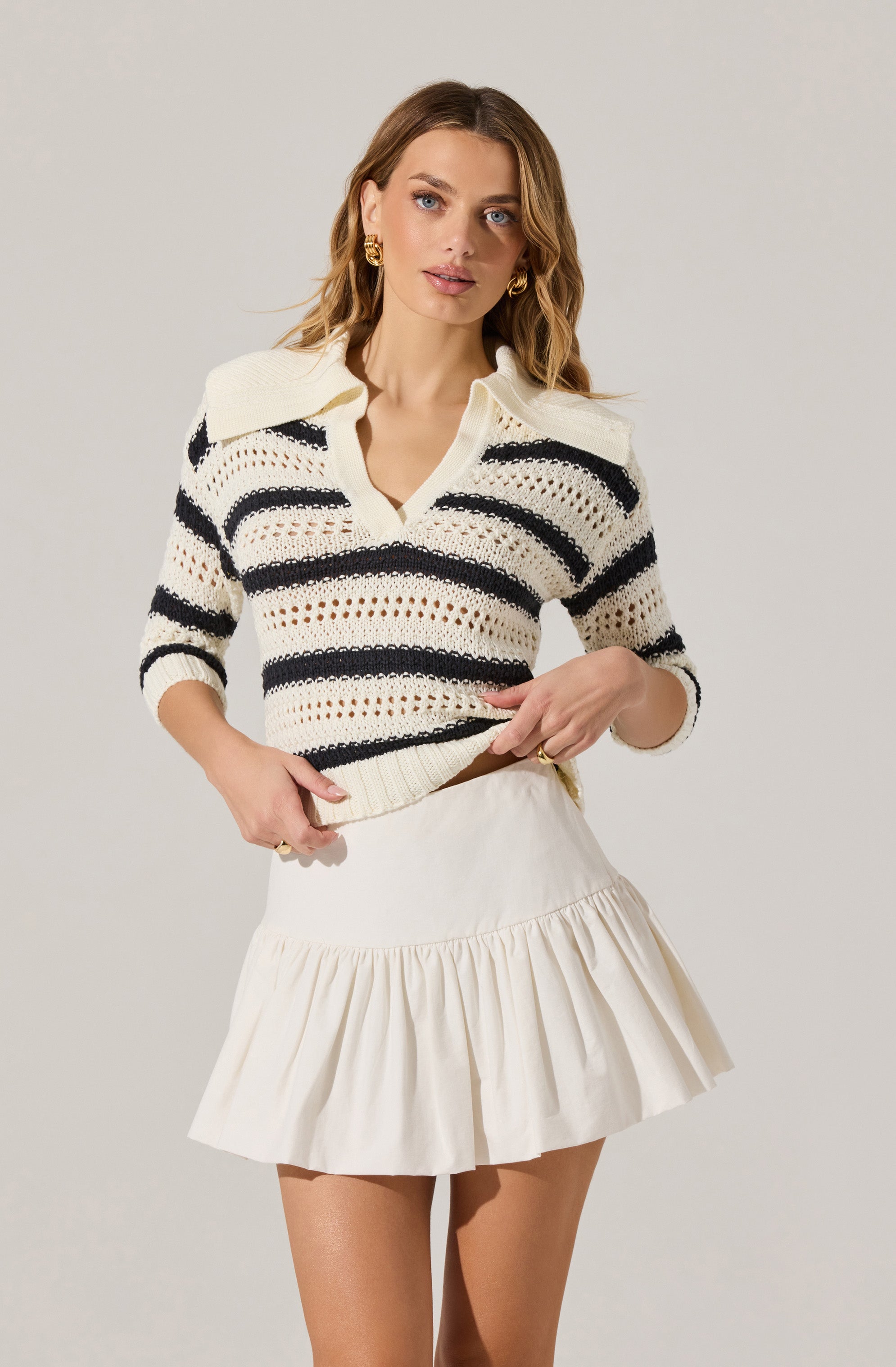 A woman is wearing a cream-colored, knit sweater with black horizontal stripes and a wide collar. She is also wearing a cream-colored, high-waisted skirt with a ruffled hem.