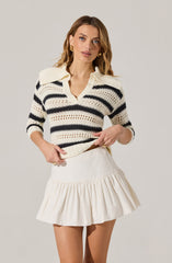 A woman is wearing a cream-colored, knit sweater with black horizontal stripes and a wide collar. She is also wearing a cream-colored, high-waisted skirt with a ruffled hem.
