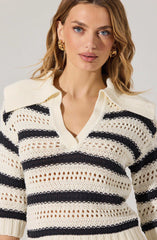 A woman is wearing a cream-colored, crochet-knit sweater with black horizontal stripes. The sweater has a V-neckline and a collar. She is accessorized with large, gold hoop earrings.