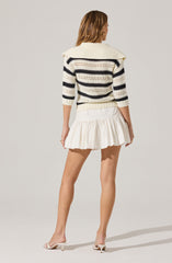 A woman is wearing a cream-colored, loose-knit sweater with wide, navy blue horizontal stripes. The sweater has a large, wide collar that drapes over her shoulders. She pairs it with a white, ruffled mini skirt that has a gathered, layered design. She is also wearing white, open-toed, heeled sandals.