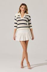 A woman is wearing a knitted sweater with horizontal black and cream stripes, featuring a fold-over collar and three-quarter length sleeves. She pairs it with a white, ruffled pleated skirt and white open-toe high-heeled sandals.