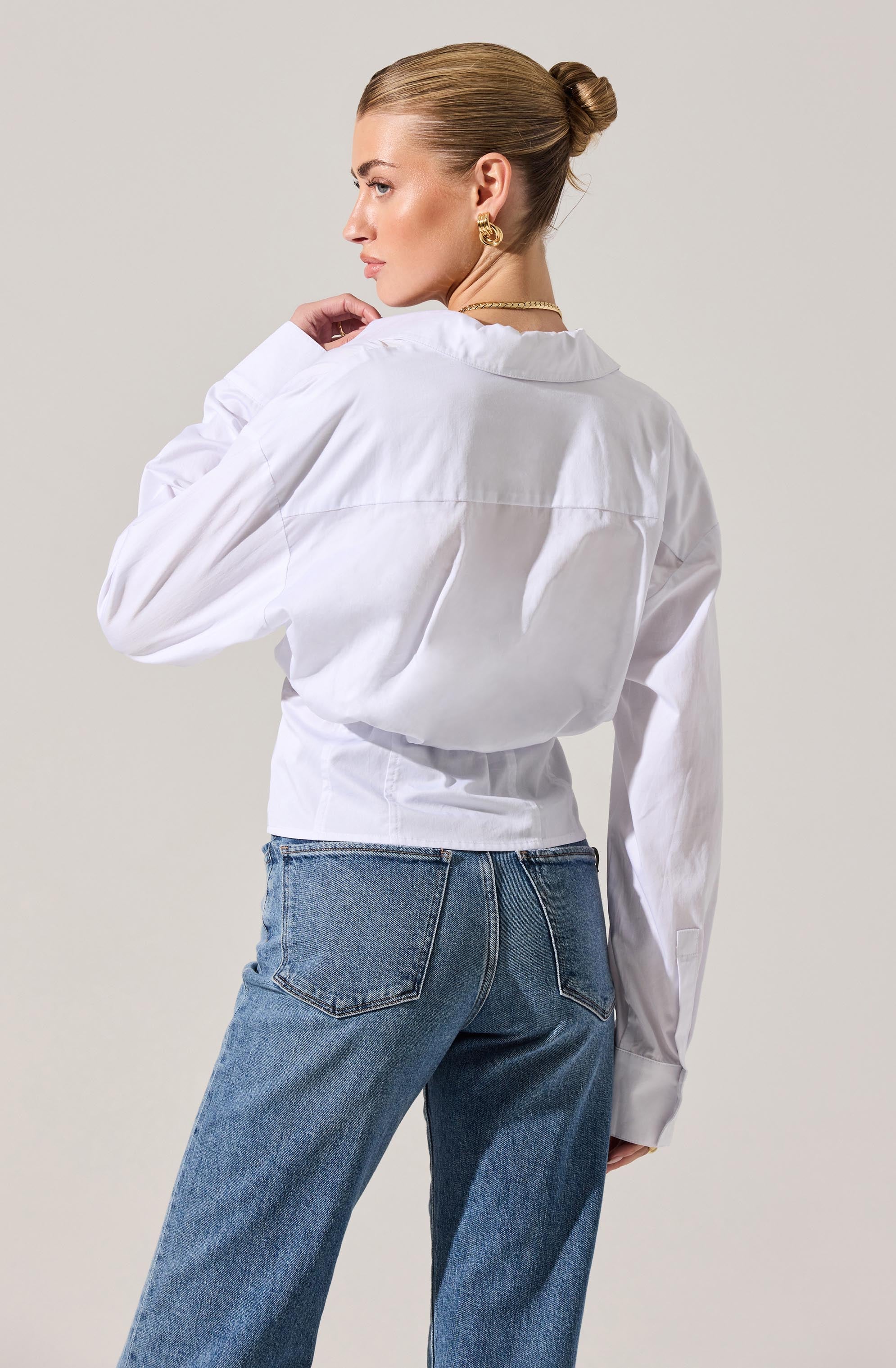 A woman is wearing a long-sleeved white button-up shirt with a collar. The shirt is slightly oversized, with a loose fit around the waist, and the sleeves are gathered at the cuffs. She pairs the shirt with high-waisted blue jeans that have a classic five-pocket design.