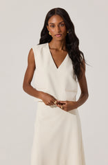 A woman is wearing a sleeveless, cream-colored dress with a V-neckline and dark brown trim along the neckline. The dress has a fitted bodice and a long, flowing skirt. She accessorizes with gold earrings and rings on her fingers, and her dark hair is styled in loose waves.