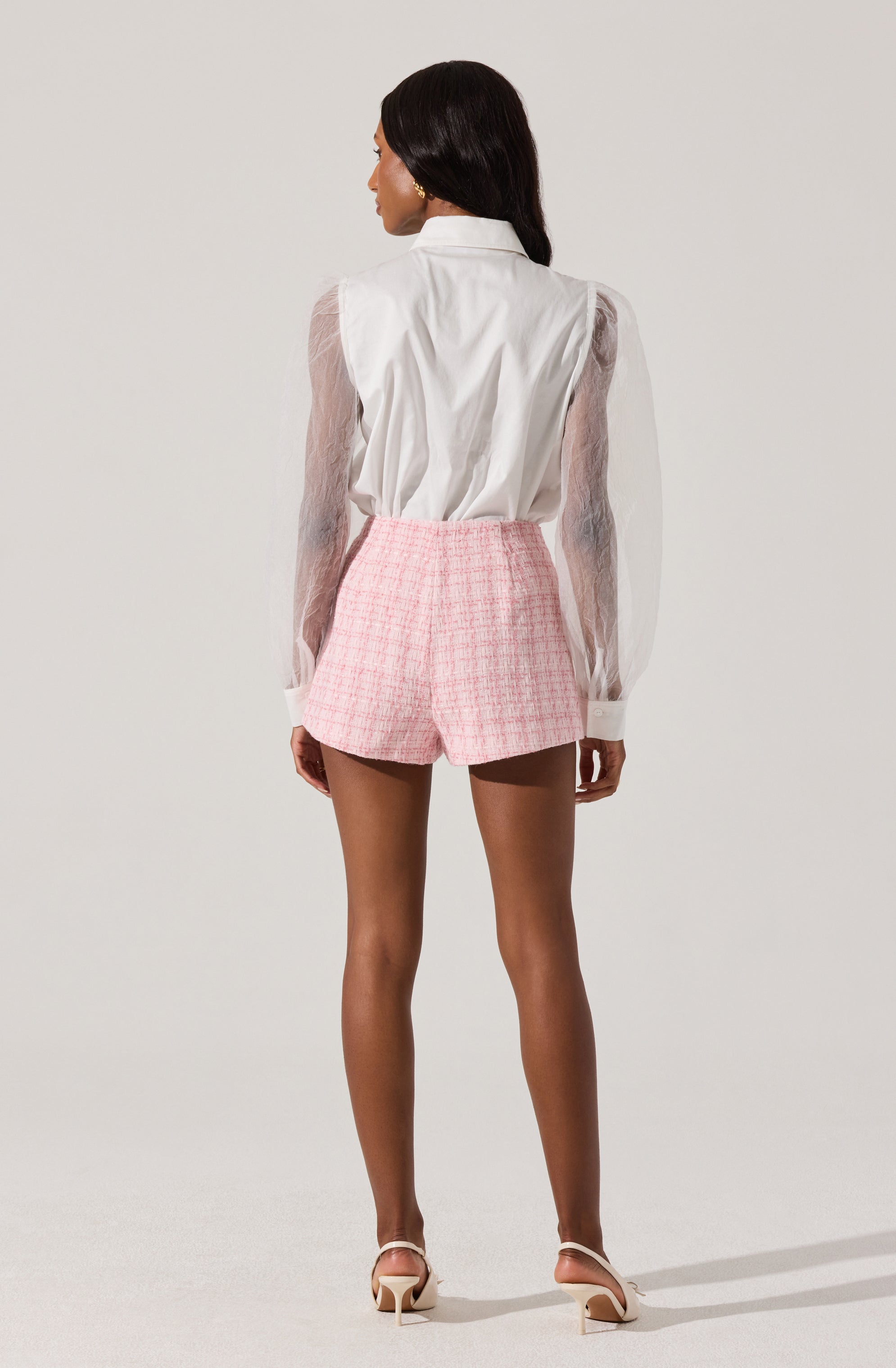 A woman is wearing a white long-sleeve blouse with sheer, slightly puffed sleeves and a collar. She pairs it with high-waisted pink plaid shorts and white high-heeled sandals.