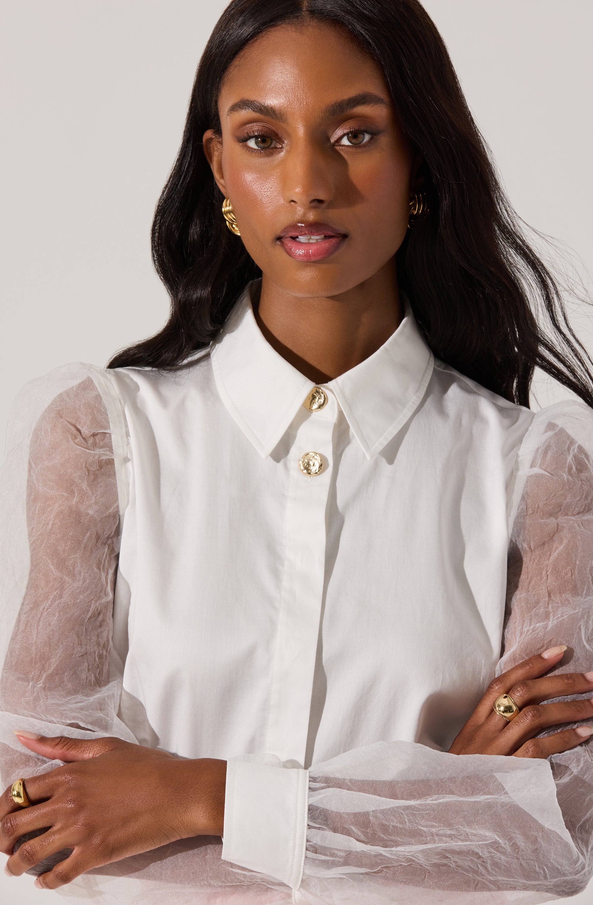A woman wearing a white button-up shirt with a pointed collar and three decorative gold buttons at the top. The shirt has sheer, puffy long sleeves made of a translucent, slightly crinkled fabric. She has gold hoop earrings, a gold ring on her left hand, and a gold ring on her right hand.