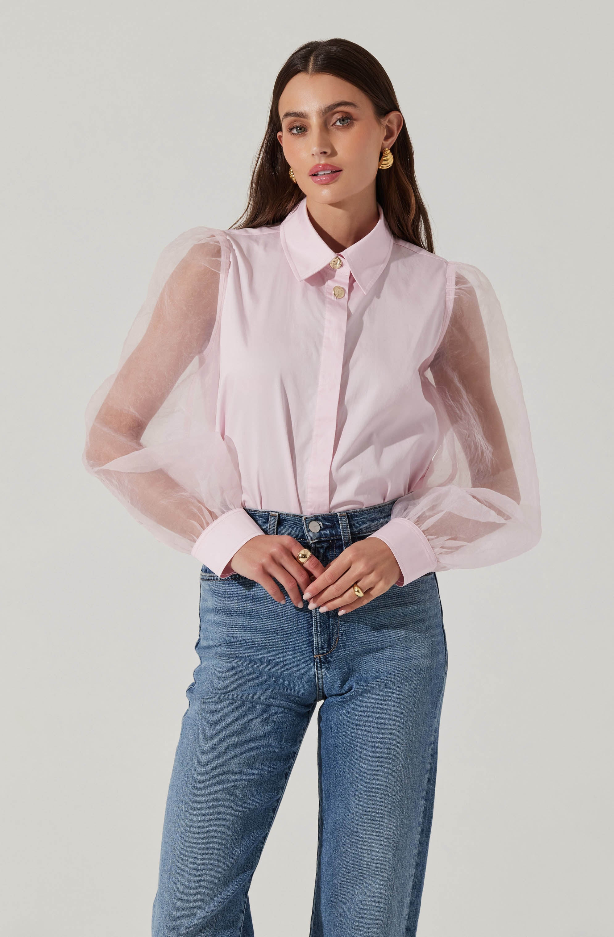 A woman is wearing a light pink button-up shirt with sheer, puffed long sleeves and a pointed collar. She has paired it with high-waisted blue jeans. She is accessorized with gold hoop earrings and rings, and her dark hair is parted in the middle.