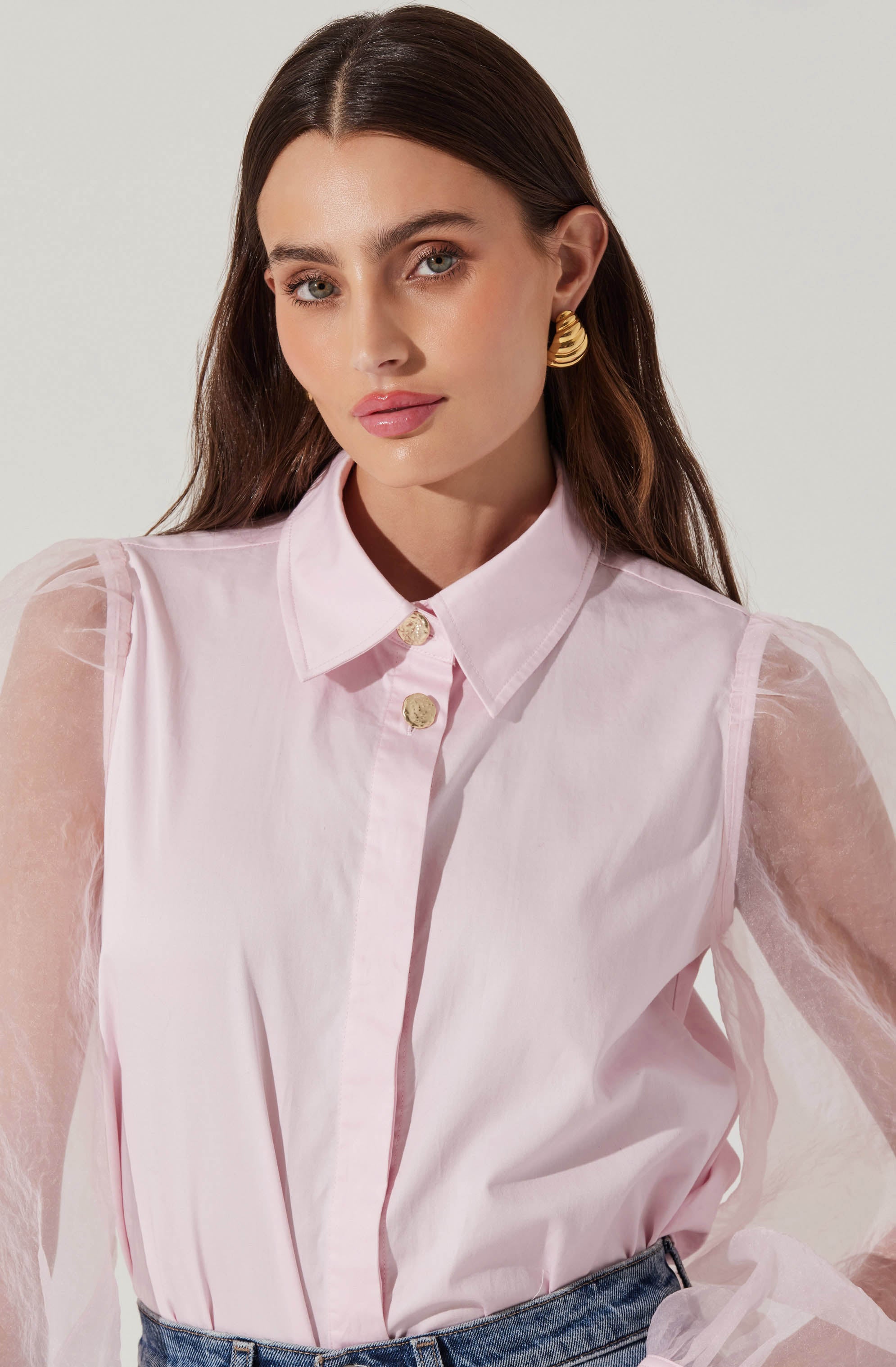 A woman is wearing a light pink button-up shirt with a classic collar. The shirt features sheer, puffy long sleeves made of a delicate, translucent fabric. She is also wearing gold statement earrings and blue jeans.