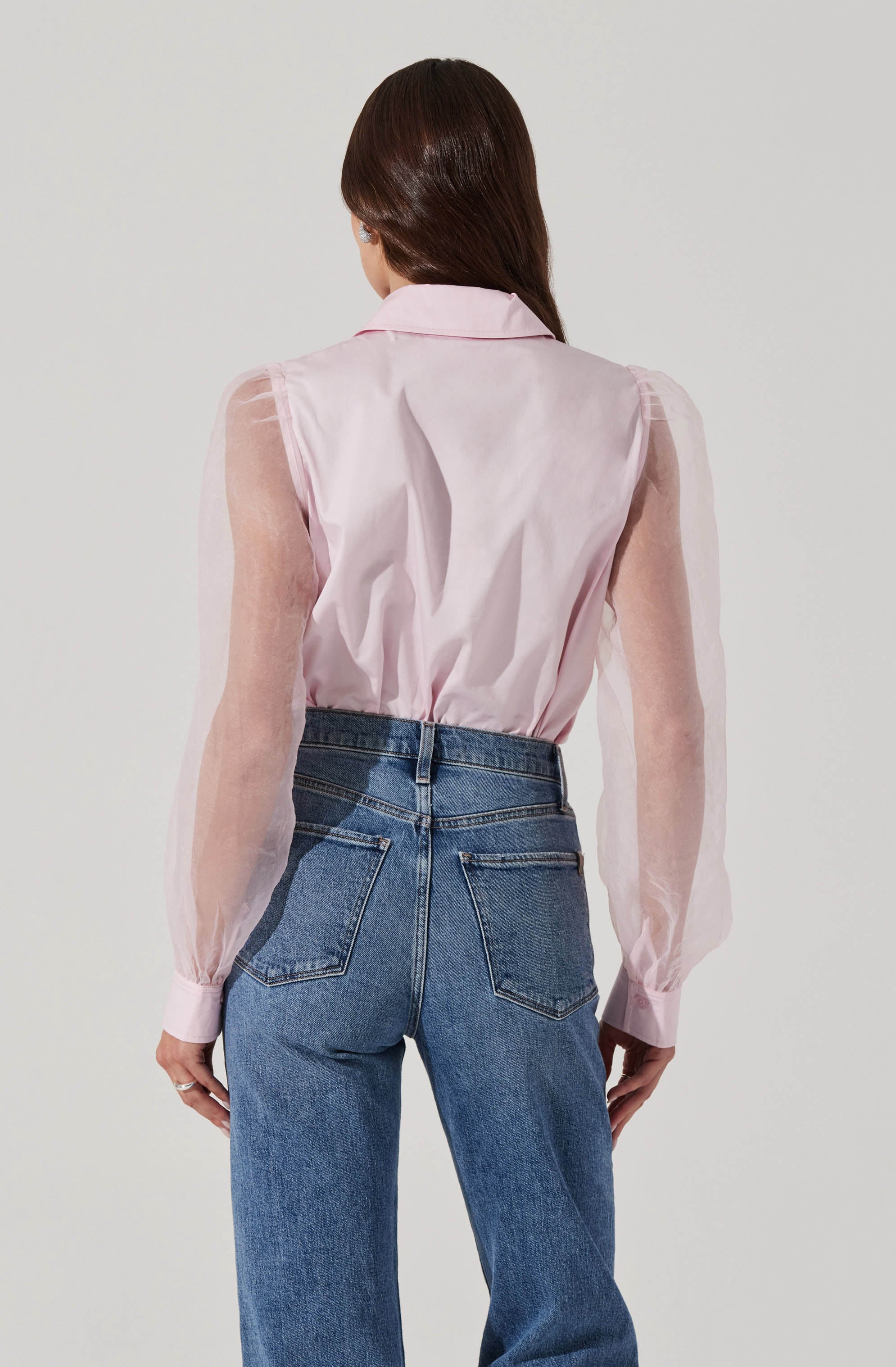 A person is wearing a sheer, light pink long-sleeve blouse with puffed sleeves and a collar, tucked into high-waisted blue jeans with back pockets.
