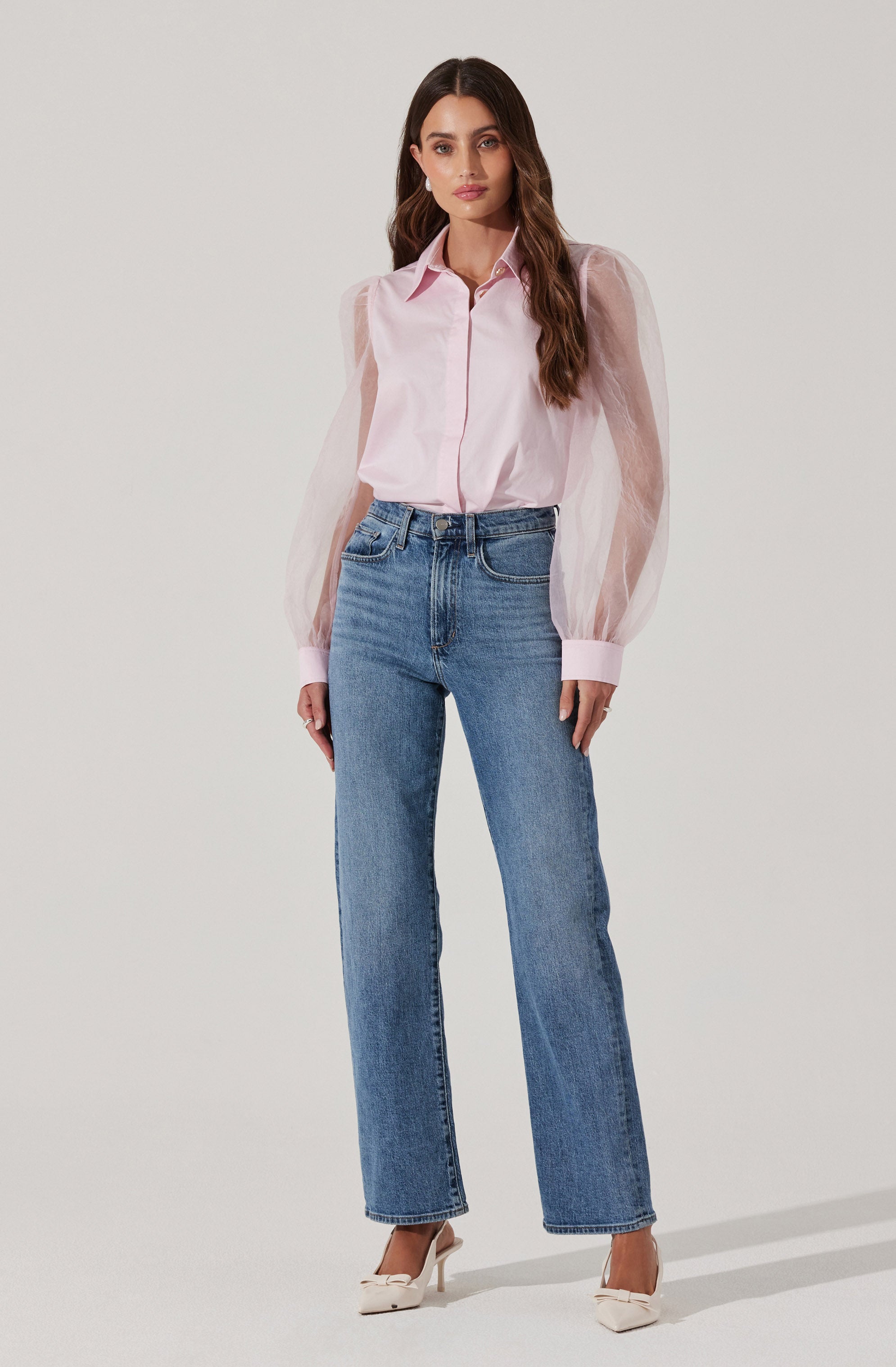 A woman is wearing a light pink, long-sleeved blouse with puffed sleeves and a collar. She has paired it with high-waisted, straight-leg blue jeans and white high-heeled shoes.