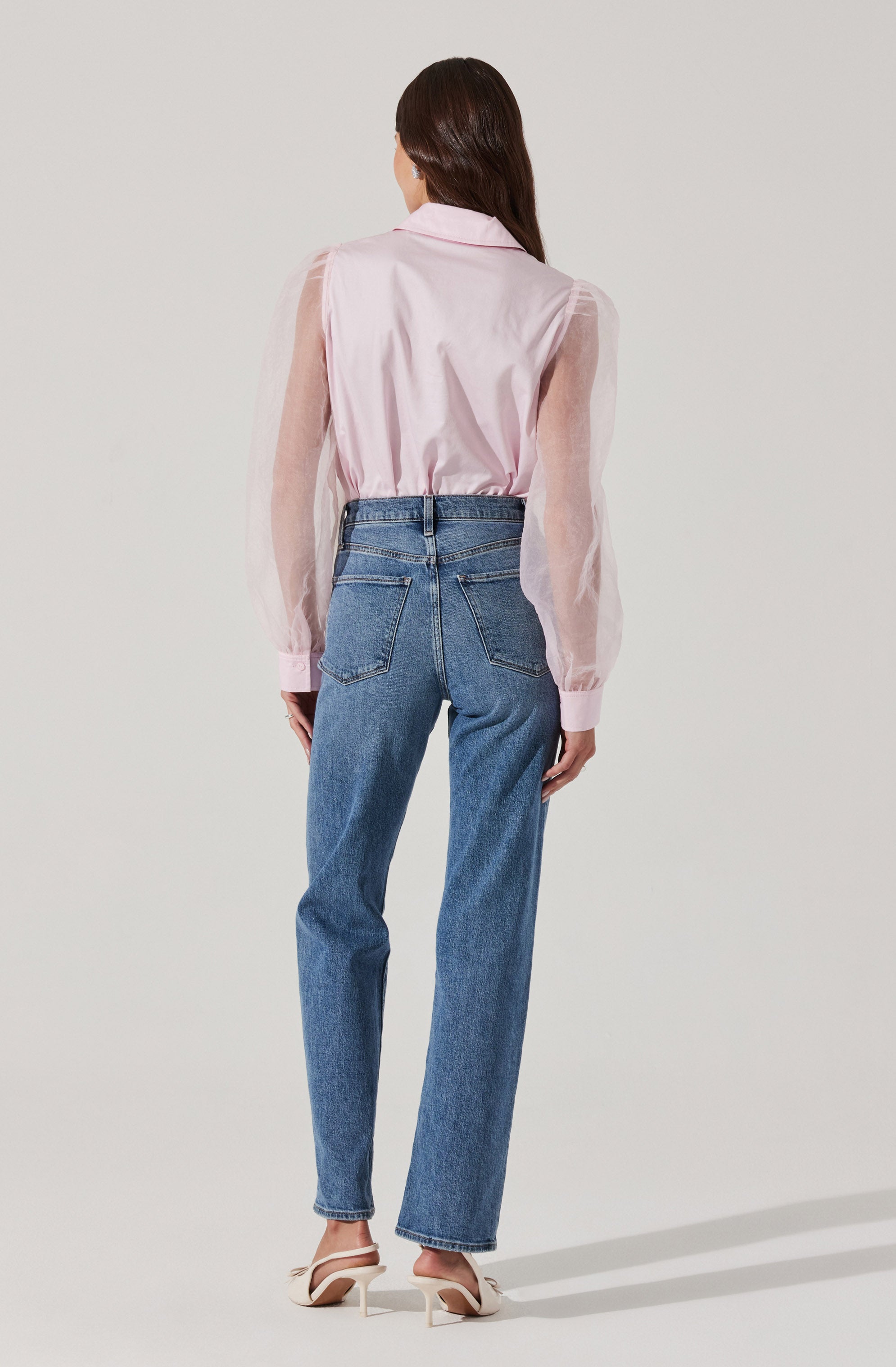 A woman is wearing a light pink, sheer, long-sleeved blouse with a collar, paired with high-waisted blue jeans that have a relaxed fit and visible back pockets. She is also wearing white heels with a pointed toe.