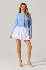 A woman is wearing a light blue, long-sleeved, cable-knit cardigan with pearl buttons down the front. She pairs the cardigan with a short, white, gathered skirt that has a puffy, voluminous shape. She is also wearing open-toed, light-colored high-heeled sandals.