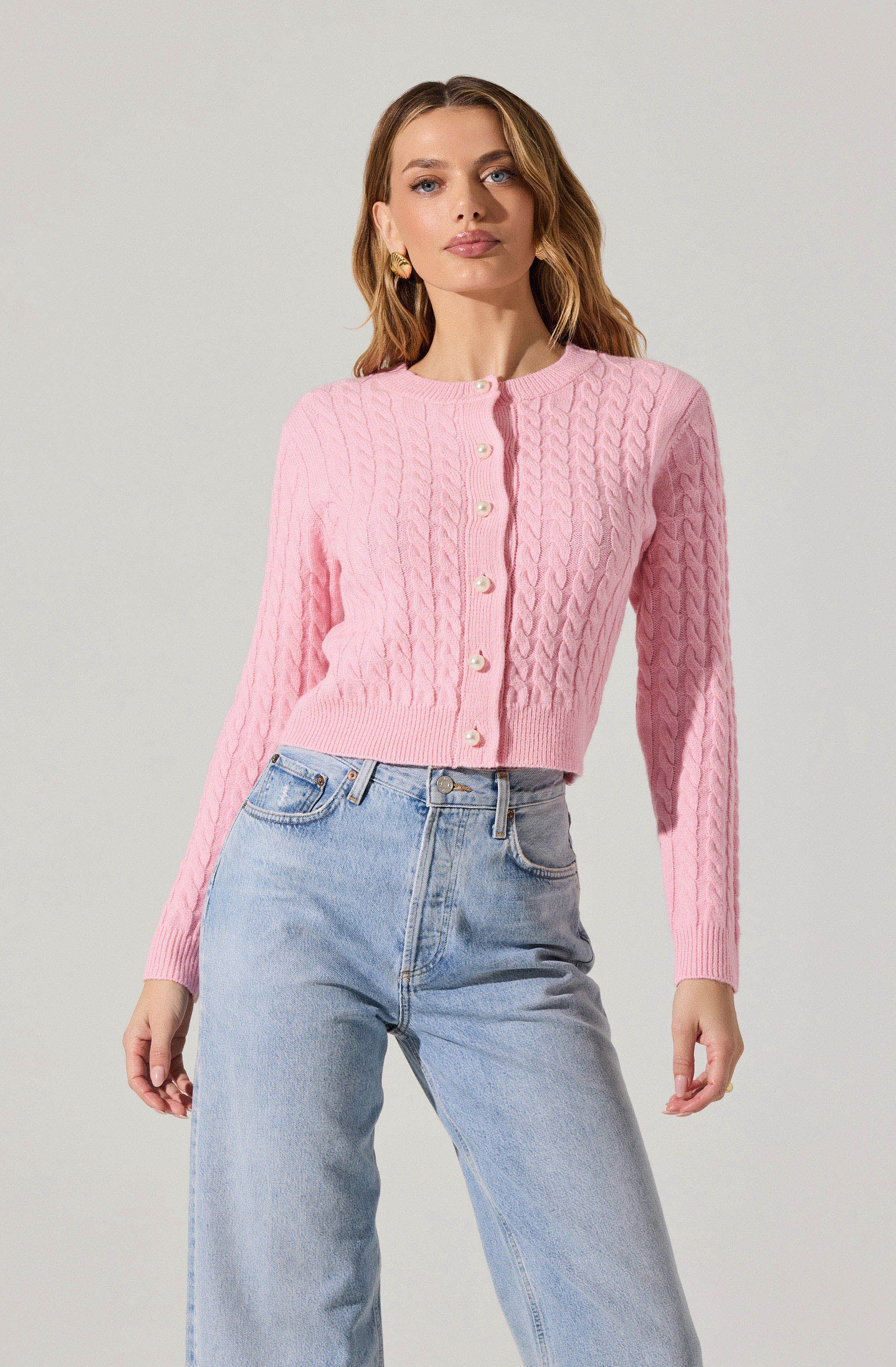 A woman is wearing a pink, long-sleeved, cable-knit cardigan with pearl buttons down the front. She is also wearing light-wash, high-waisted jeans.