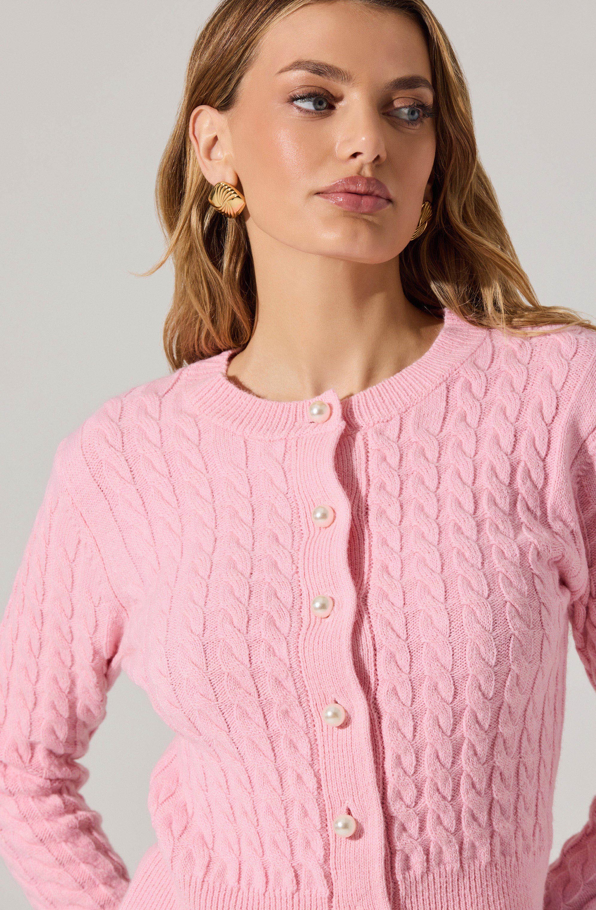 A woman is wearing a light pink, cable-knit cardigan with a round neckline, featuring pearl buttons down the front. She has gold shell-shaped earrings and her hair is styled in loose waves.
