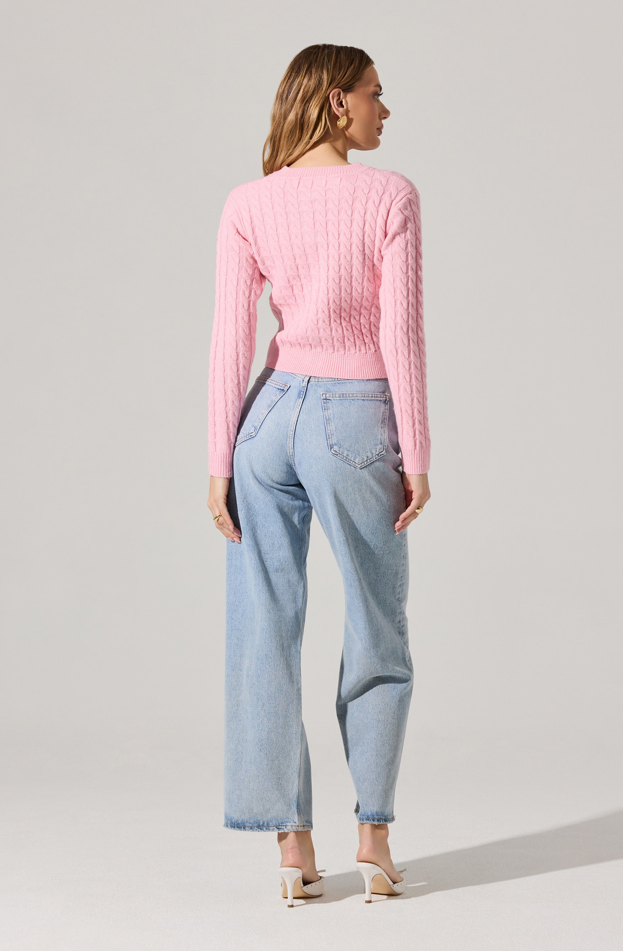 A woman is wearing a pink, cable-knit sweater with long sleeves and a ribbed hem. She pairs it with high-waisted, light-wash, relaxed-fit jeans that have a slightly tapered leg. She completes the look with beige high-heeled sandals.