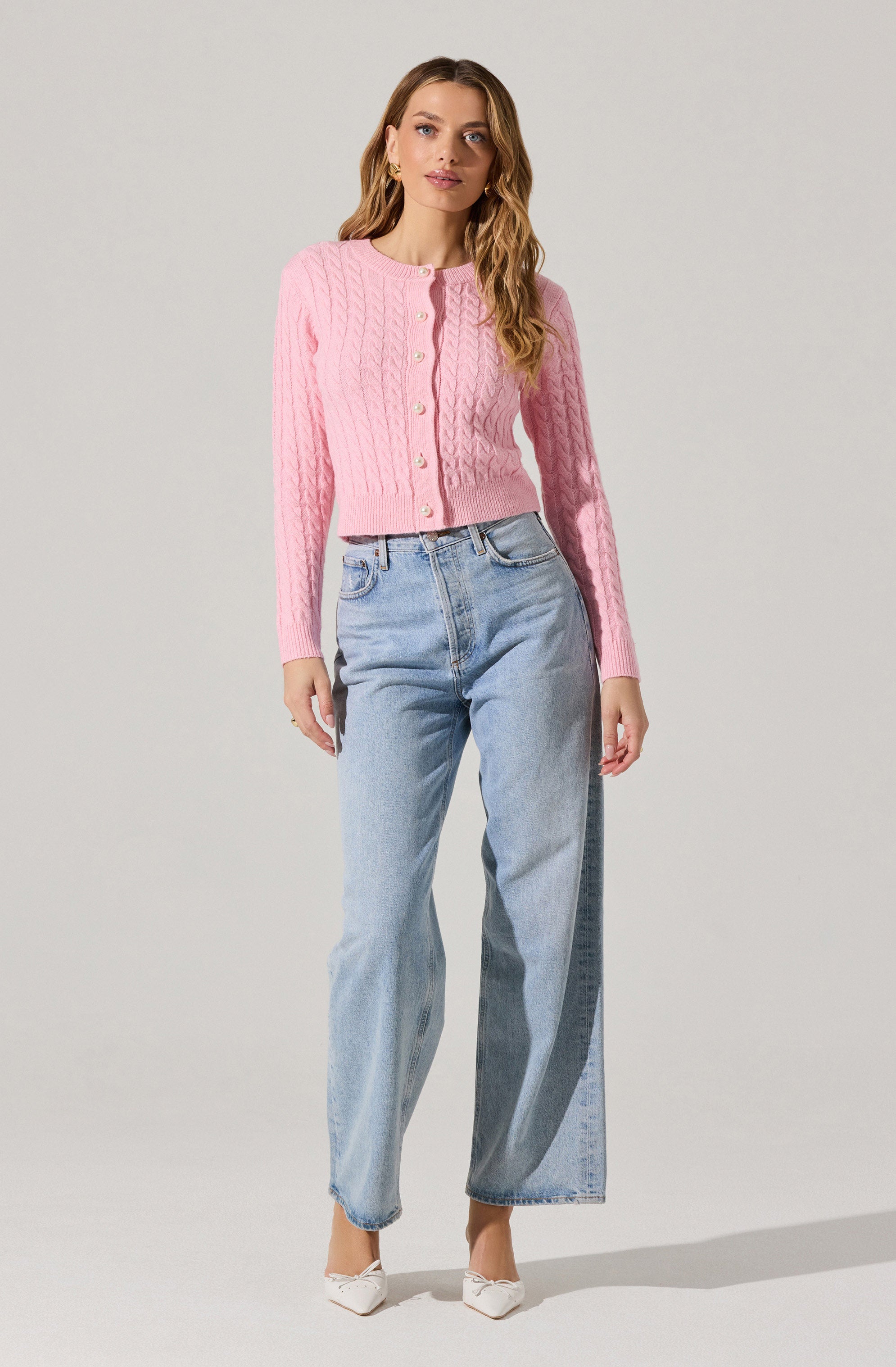 A woman is wearing a pink knit cardigan with a cable pattern and pearl buttons down the front. She pairs it with light blue high-waisted, wide-leg jeans and white pointed-toe shoes.