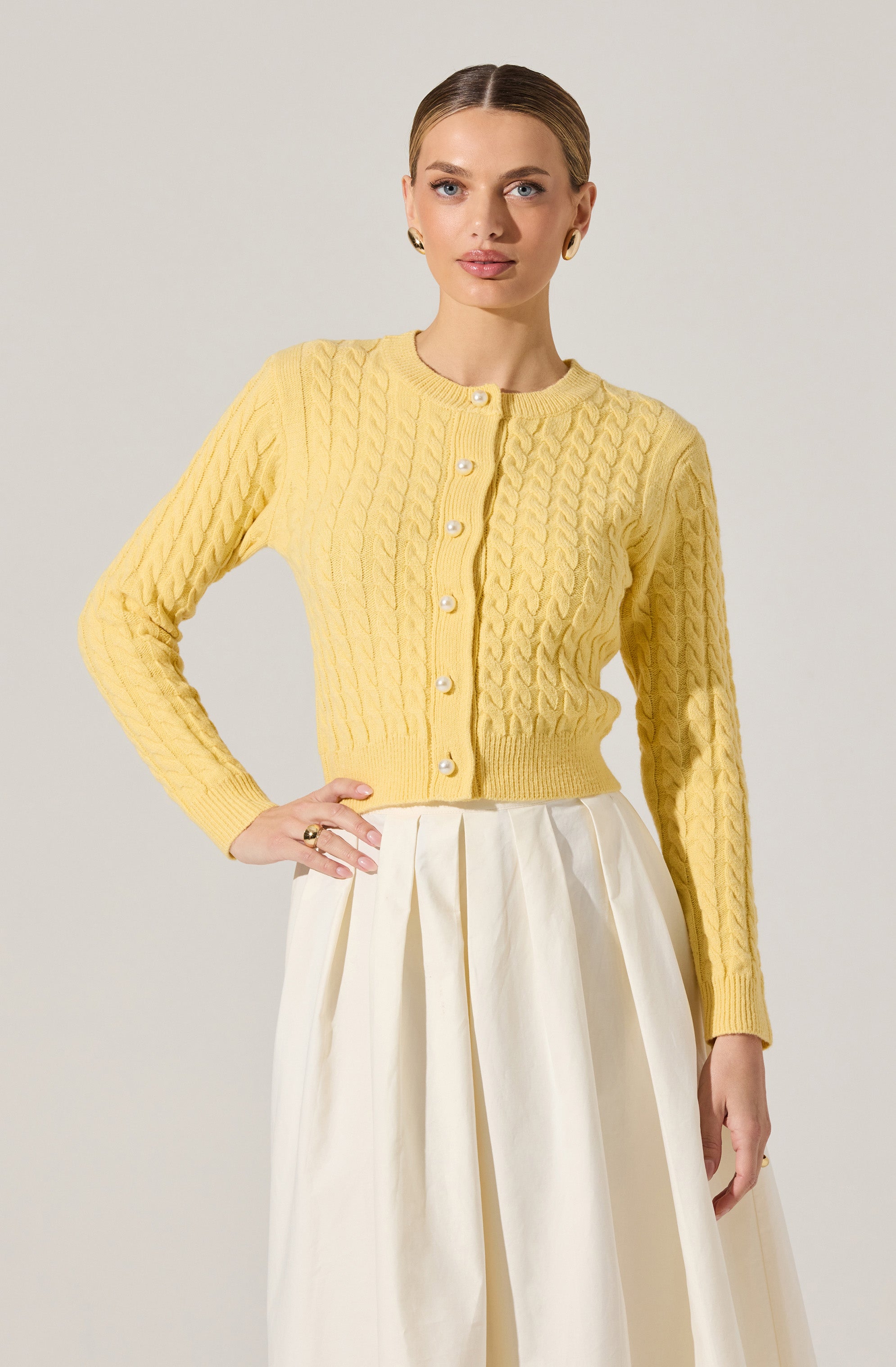 A woman is wearing a yellow, long-sleeved knitted cardigan with a cable pattern and pearl buttons down the front. She is also wearing a high-waisted, wide-legged white skirt with pleats.