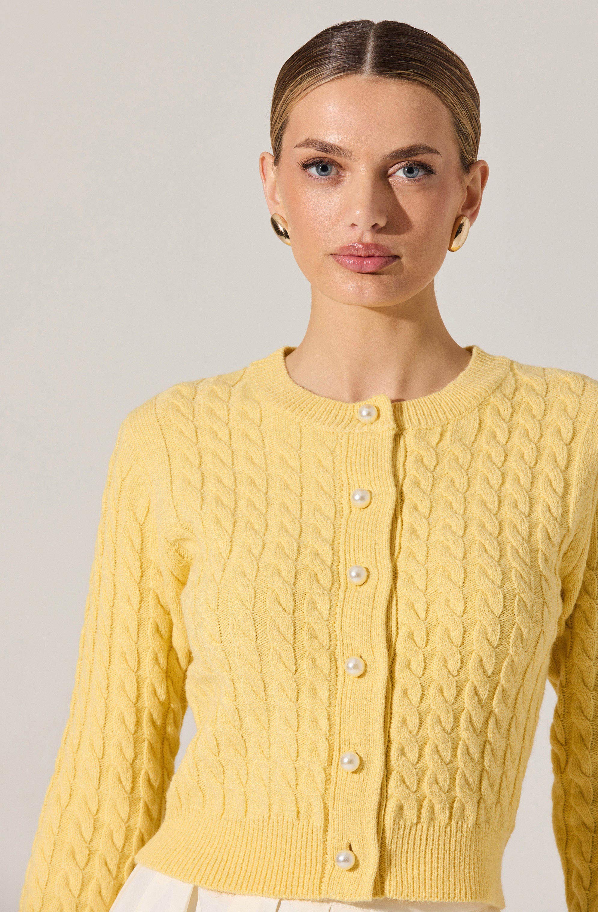 A woman is wearing a yellow knitted cardigan with a cable pattern, featuring a round neckline and pearl buttons down the front.