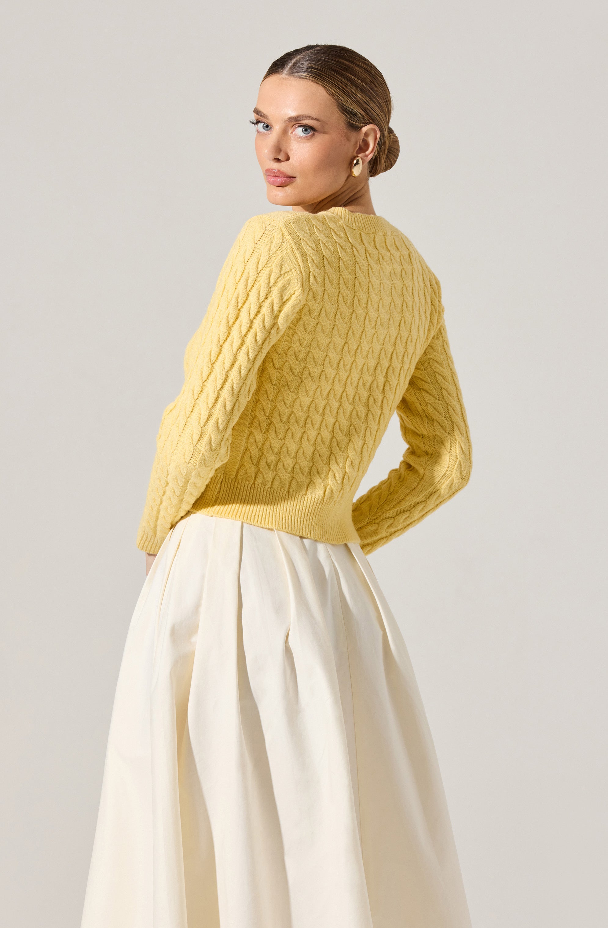 A woman is wearing a yellow cable-knit sweater with a textured pattern, featuring long sleeves and a ribbed hem. She pairs it with a high-waisted, cream-colored, pleated skirt.