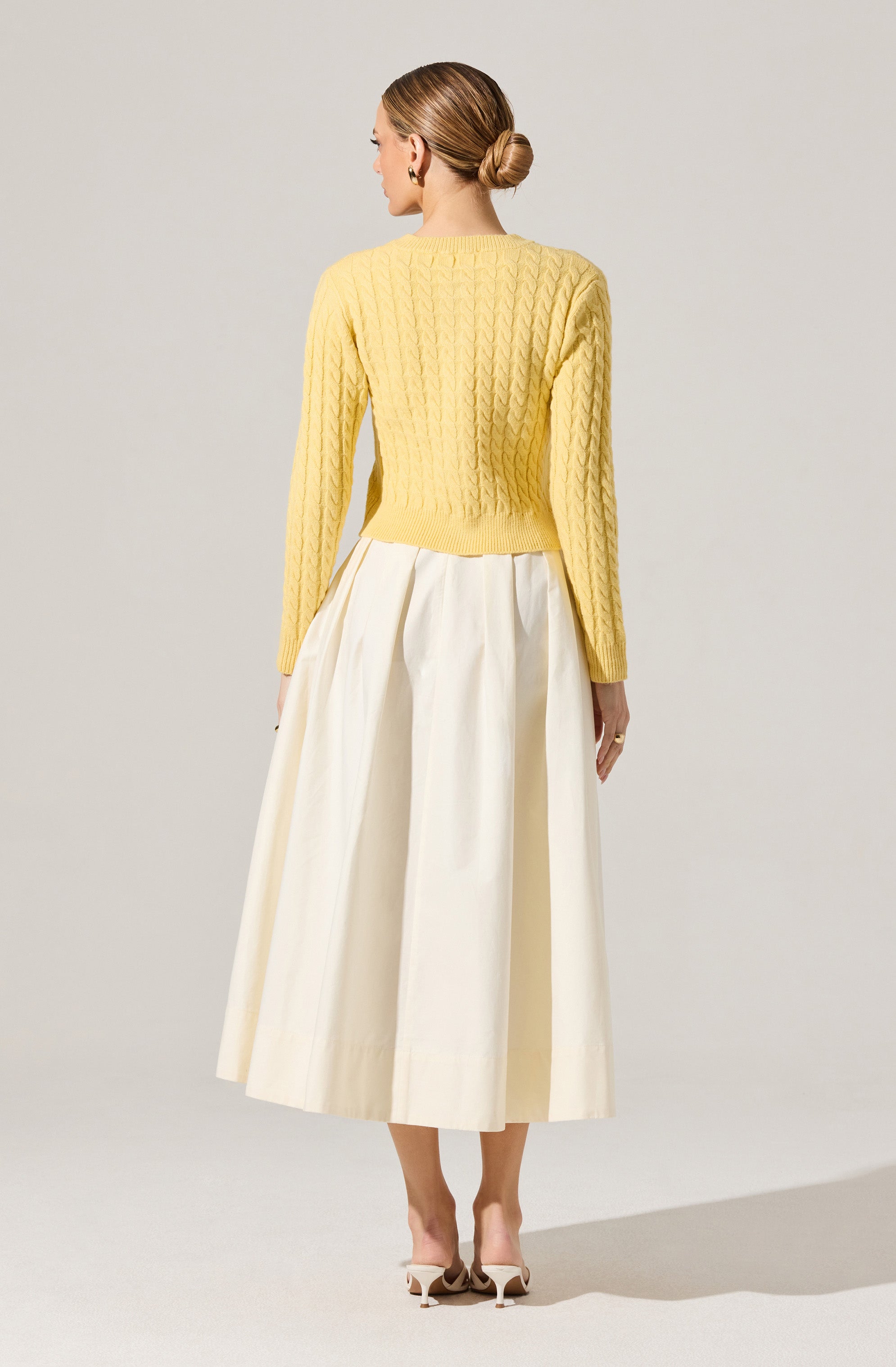A woman is wearing a bright yellow, long-sleeved, cable-knit sweater with a crew neckline, paired with an elegant, high-waisted, cream-colored, pleated midi skirt. She is also wearing light-colored heels.