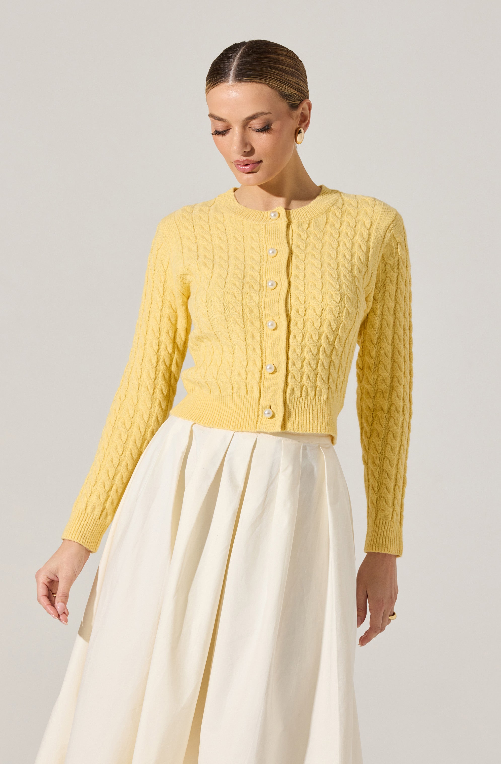 A woman is wearing a light yellow, long-sleeved, cable-knit cardigan with a round neckline and pearl buttons down the front. She has paired it with a high-waisted, cream-colored, pleated skirt.