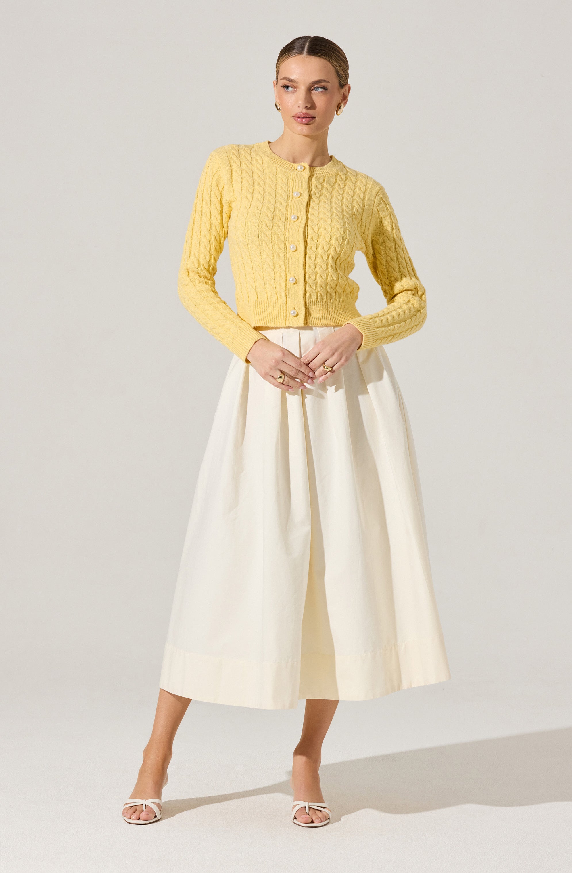 A woman is wearing a yellow knitted cardigan with a cable knit pattern and buttons down the front. She pairs it with high-waisted, wide-leg cream-colored trousers that reach mid-calf. She completes the outfit with white open-toe heels.