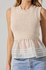 A woman is wearing a sleeveless knit top with a round neckline in a light beige color. The top has a textured, open-knit pattern running vertically down the front, with a ribbed band at the waist. The bottom of the top features a layered, sheer, cream-colored fabric with horizontal stripes, creating a peplum-like effect. She is also wearing light blue denim jeans.