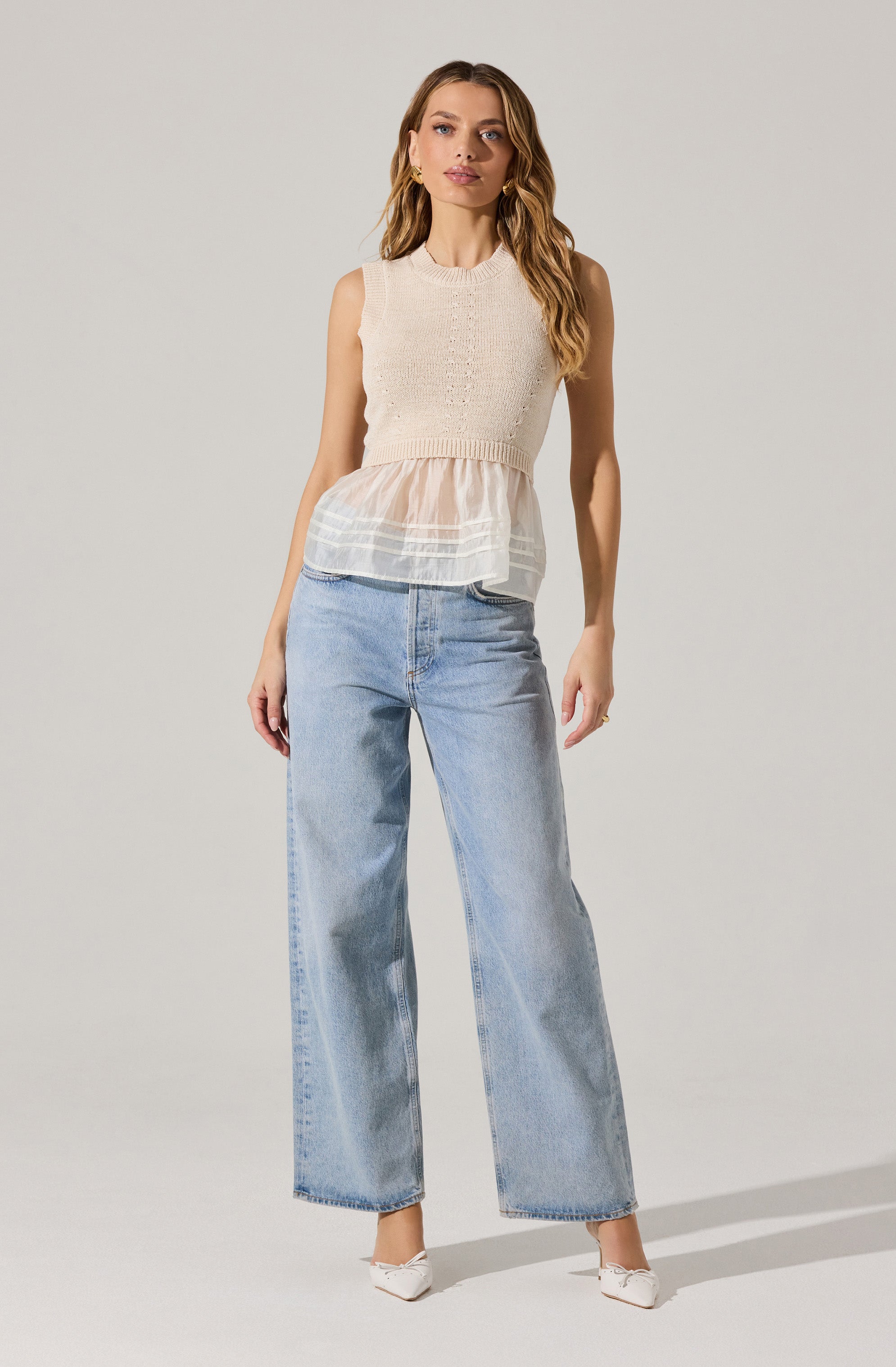 A woman is wearing a sleeveless, knitted, cream-colored top with a layered, sheer, peach-colored fabric detail at the waist. She pairs it with high-waisted, light-wash, wide-leg jeans and white pointed-toe shoes.