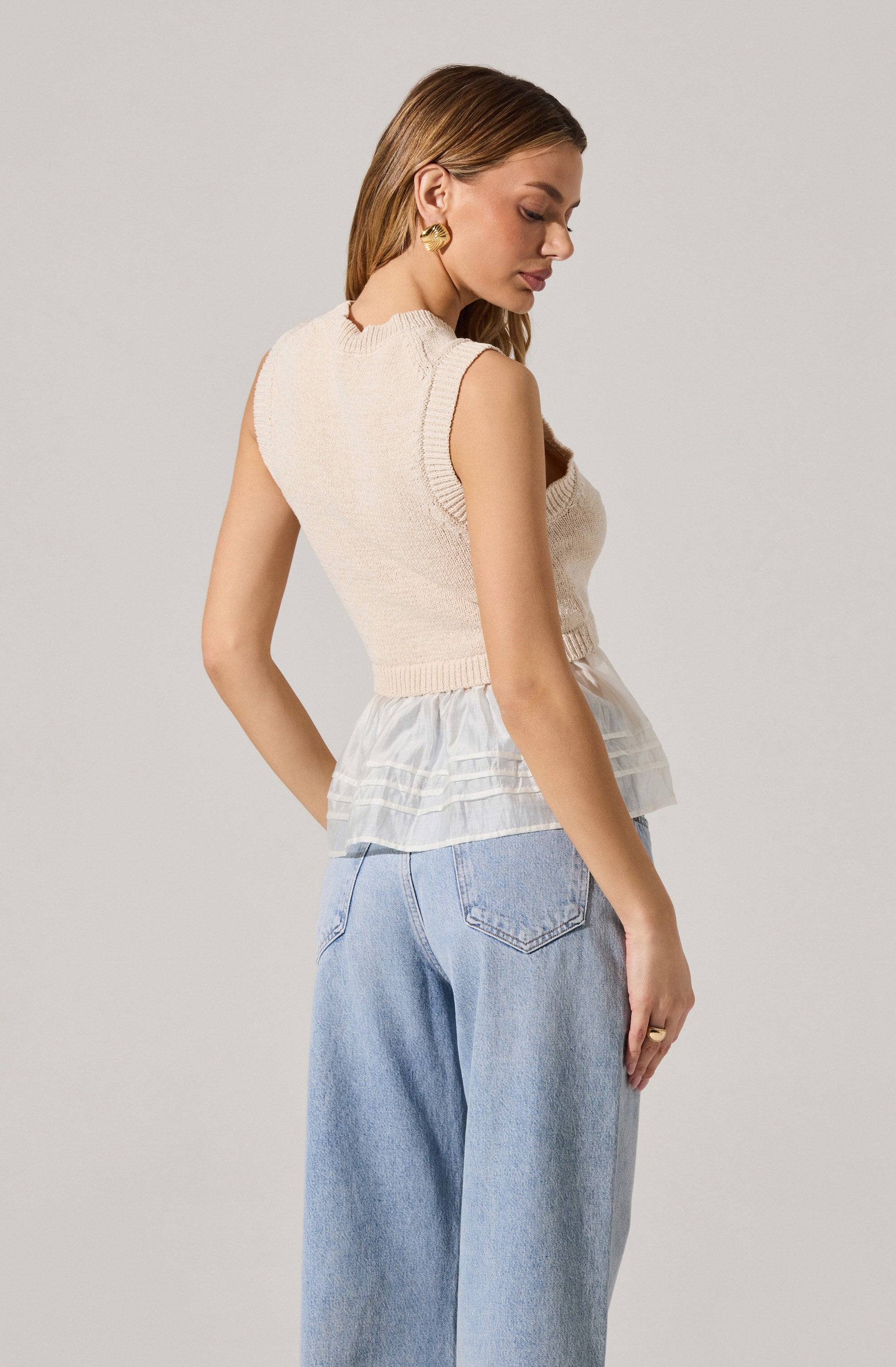 A woman is wearing a sleeveless, cream-colored knit vest with a round neckline, layered over a sheer, ruffled white blouse. She has paired the top with high-waisted, light-wash denim jeans. She is accessorized with gold hoop earrings and a ring on her left hand.