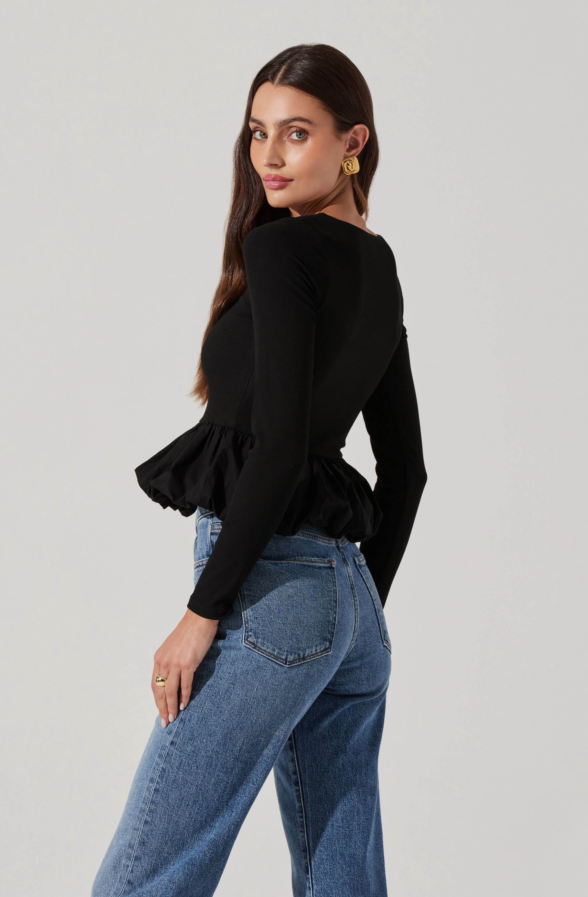 A woman is wearing a fitted black long-sleeve top with a ruffled peplum hem at the waist, paired with high-waisted blue jeans. The top has a smooth, slightly shiny fabric, and the jeans are a classic denim wash with a relaxed fit. She has long, dark hair and is accessorized with gold earrings.