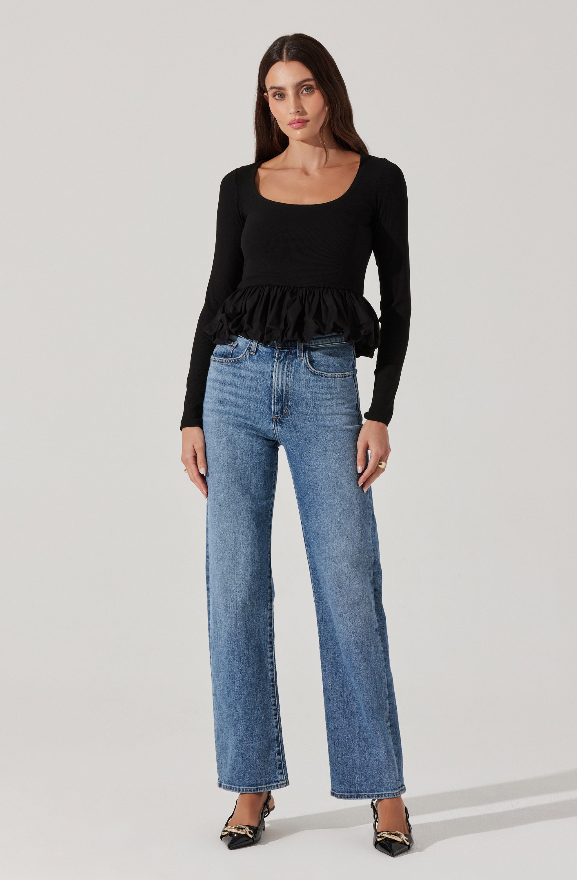 A woman is wearing a black long-sleeve top with a rounded neckline and a ruffled hemline. She pairs it with high-waisted, straight-leg blue jeans and black pointed-toe heels with embellishments.