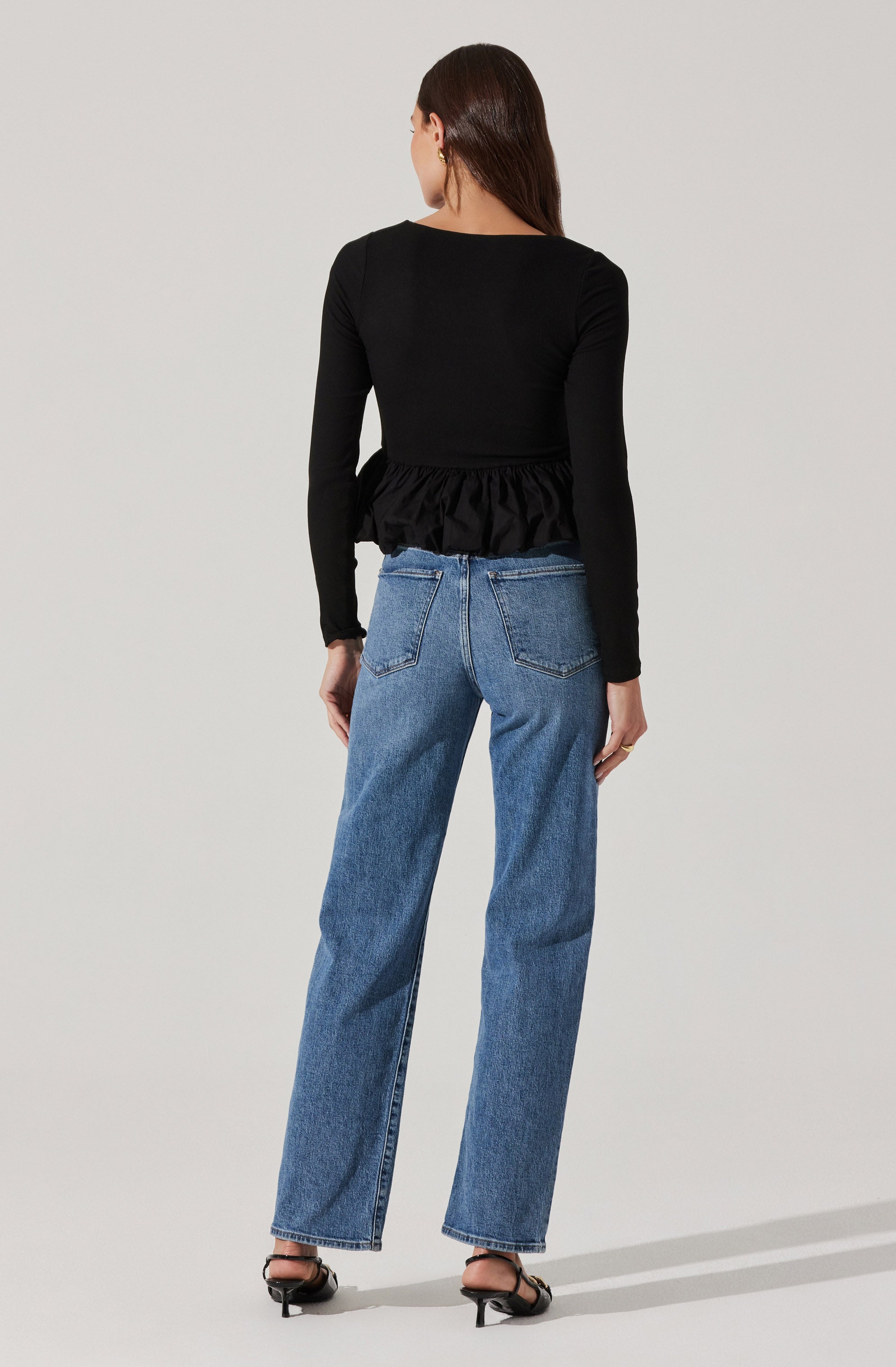 A woman is wearing a black long-sleeve top with a ruffled hem at the waist, paired with high-waisted, straight-leg blue jeans. She is also wearing black open-toe sandals with a small heel.