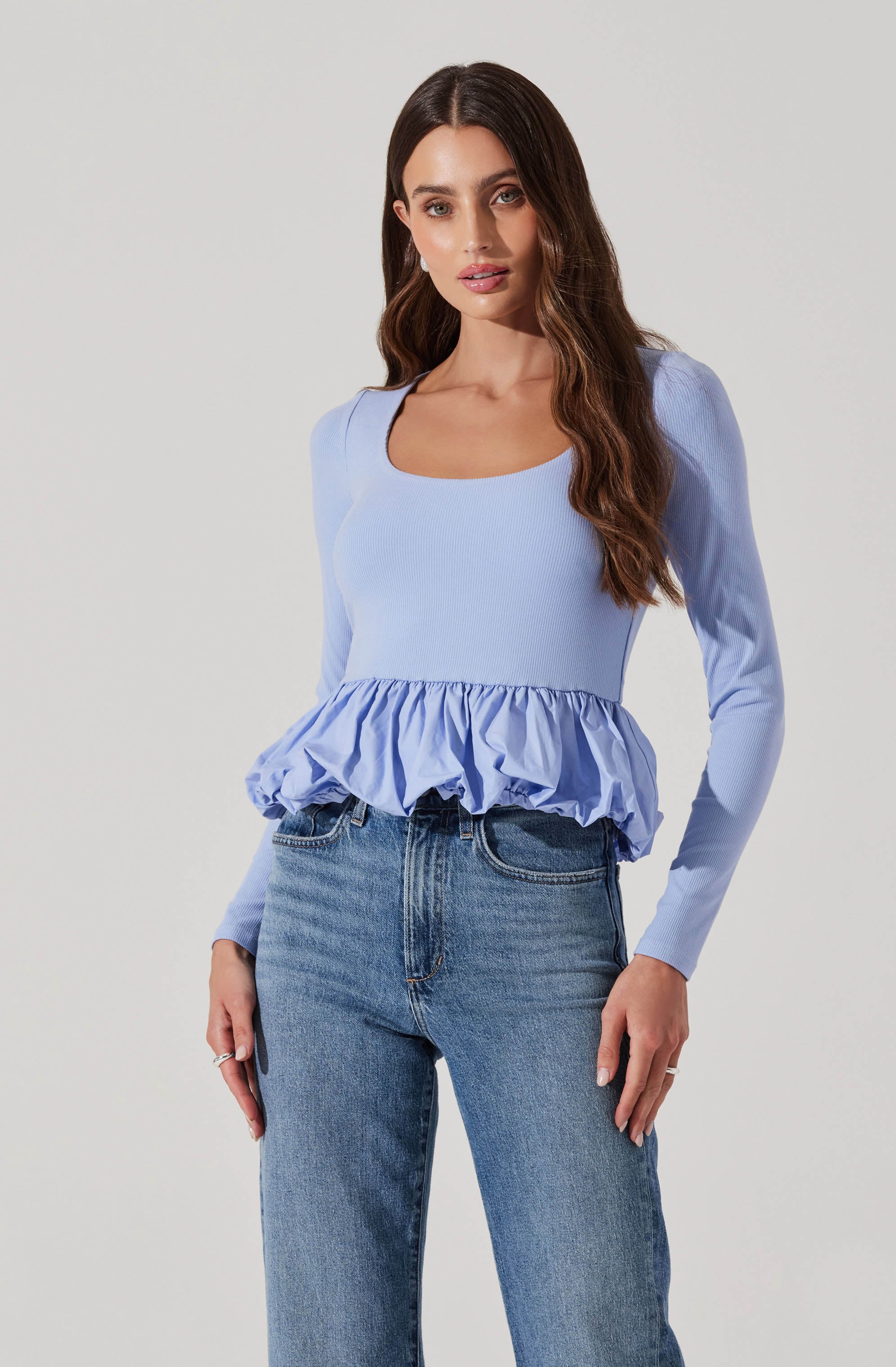 A woman is wearing a light blue long-sleeve top with a scoop neckline and a ruffled, gathered hem. She is also wearing mid-rise, straight-leg blue jeans.